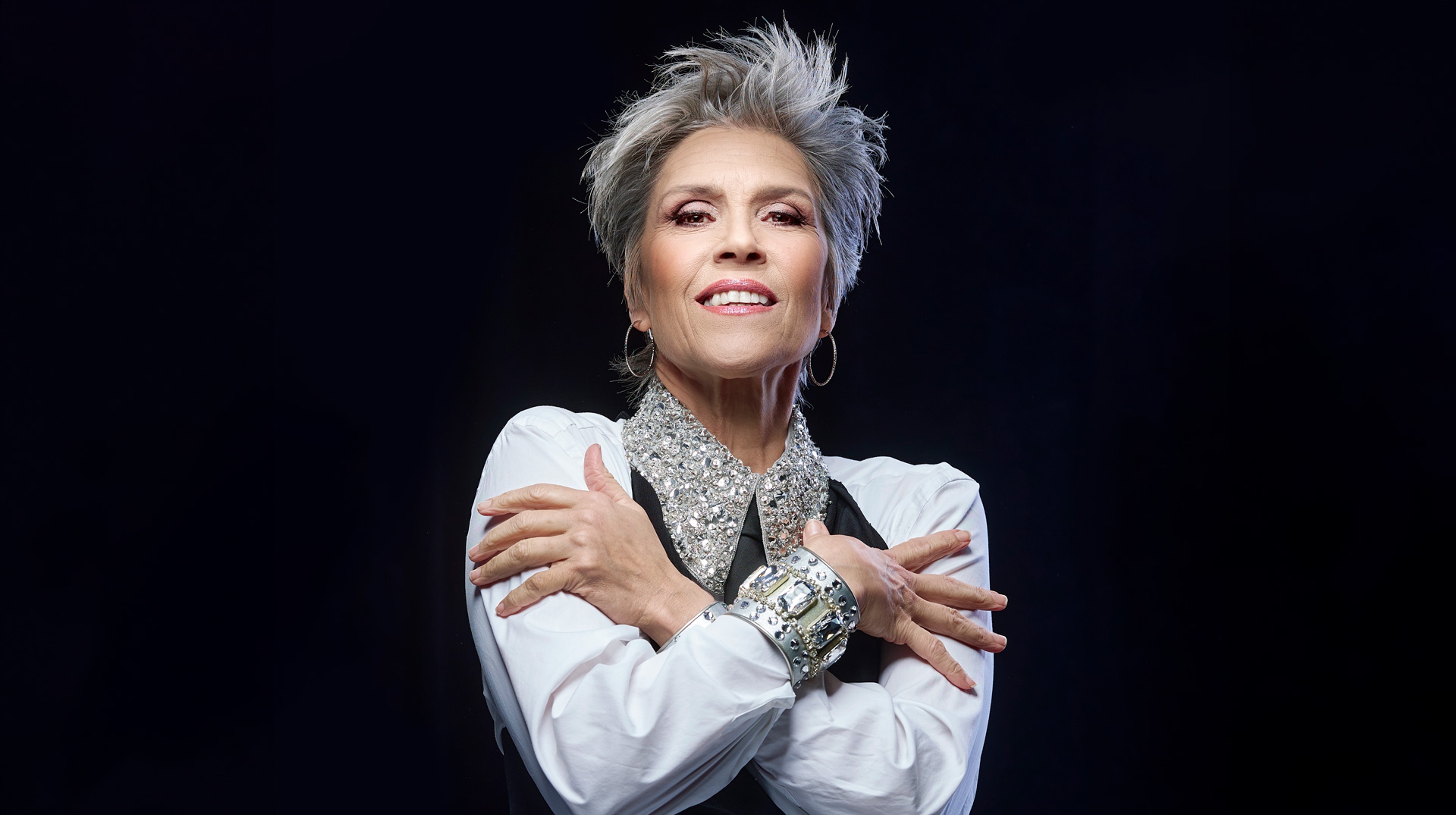 Petra Nielsen står med armarna i kors framför en svart bakgrund och ler in i kameran