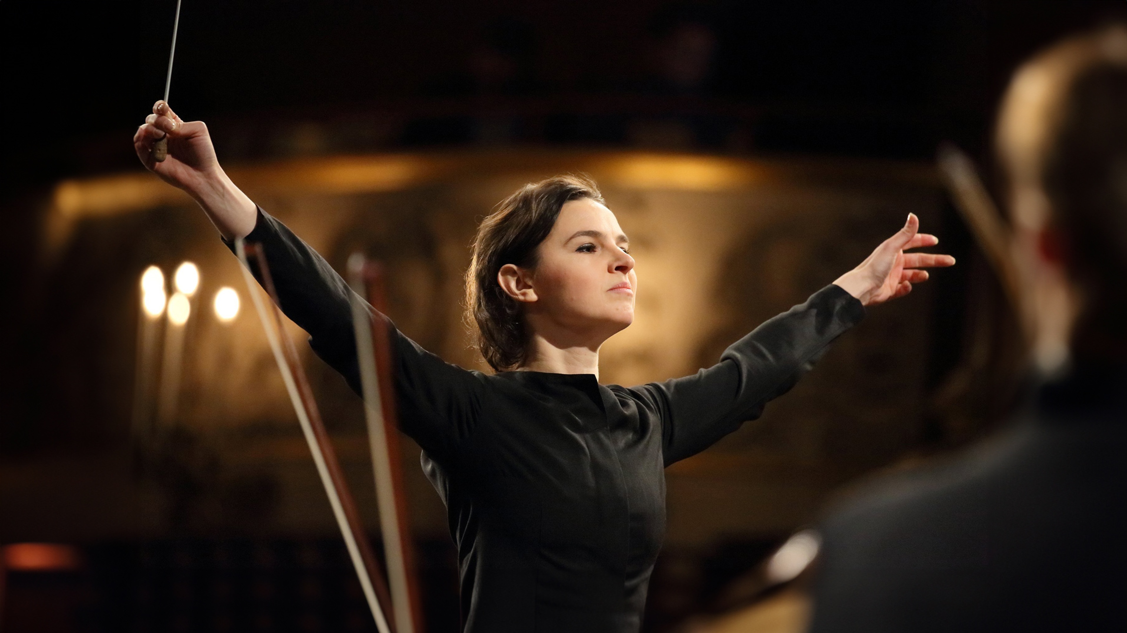 Oksana Lynnig står med händer utsträckta och dirigentpinnen i hand
