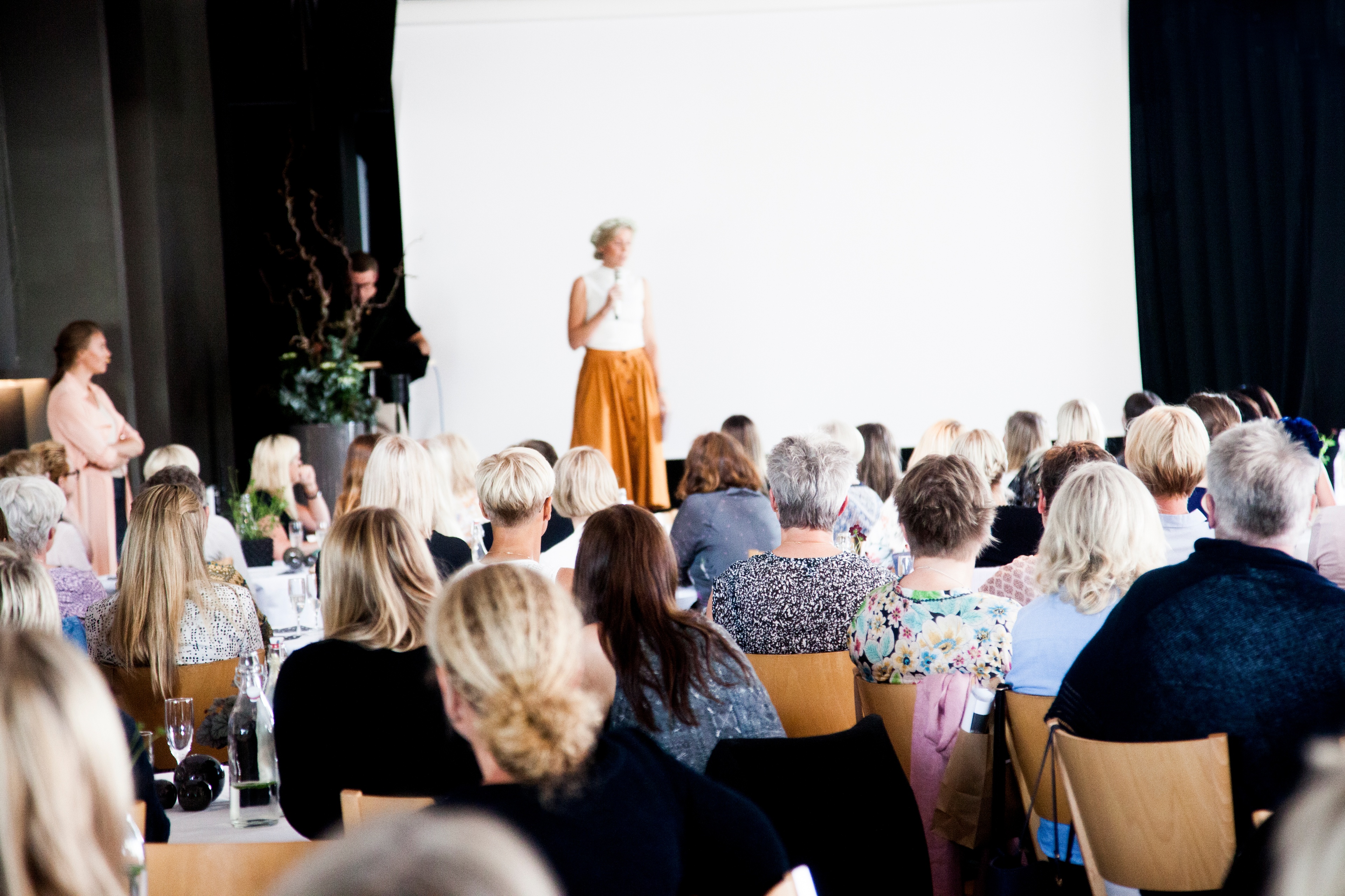 En föreläsare står på scen framför publik som sitter på stolar vid restaurangbord.