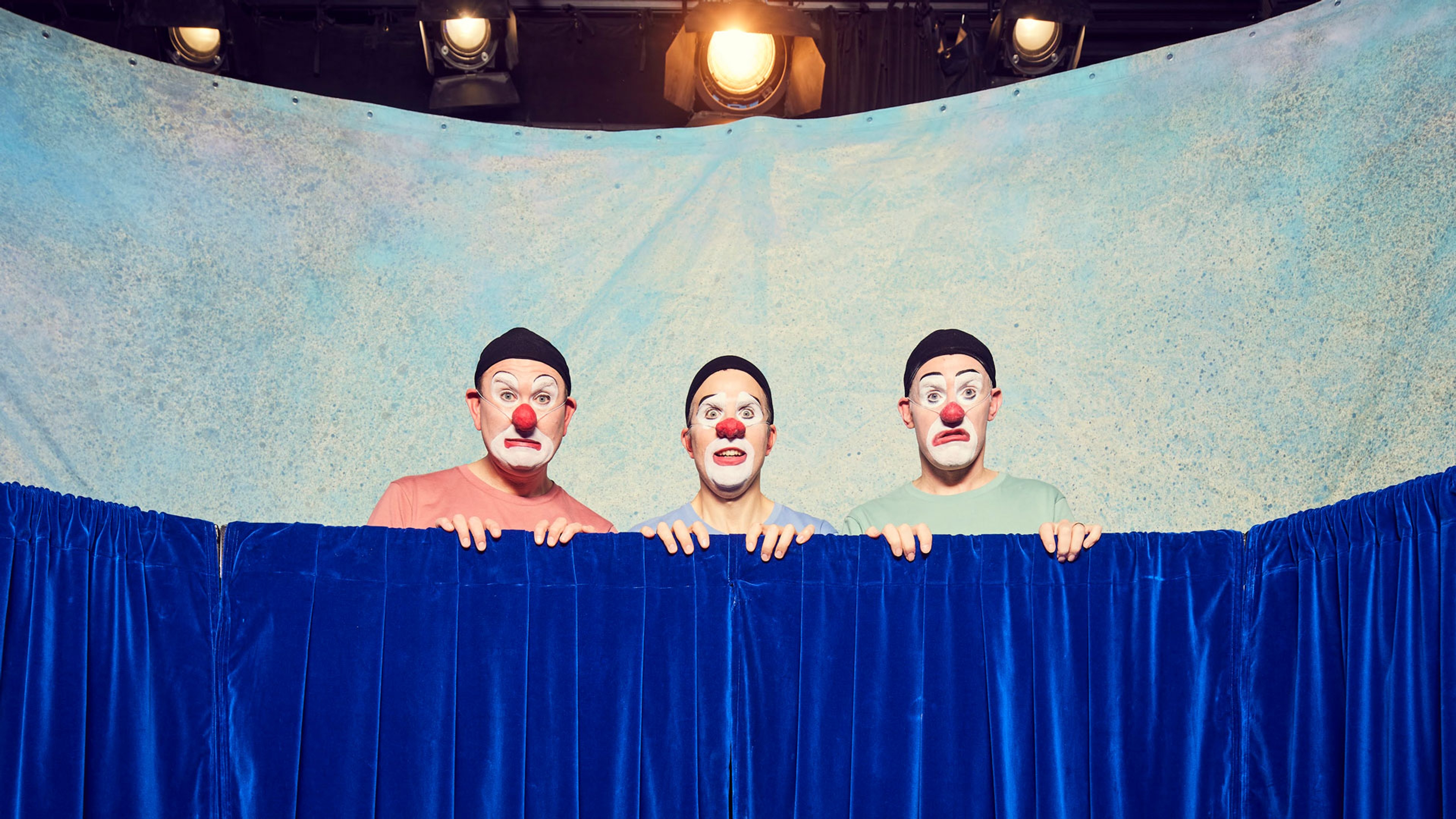 Tre personer i clownsminkning tittar upp över ett skynke.