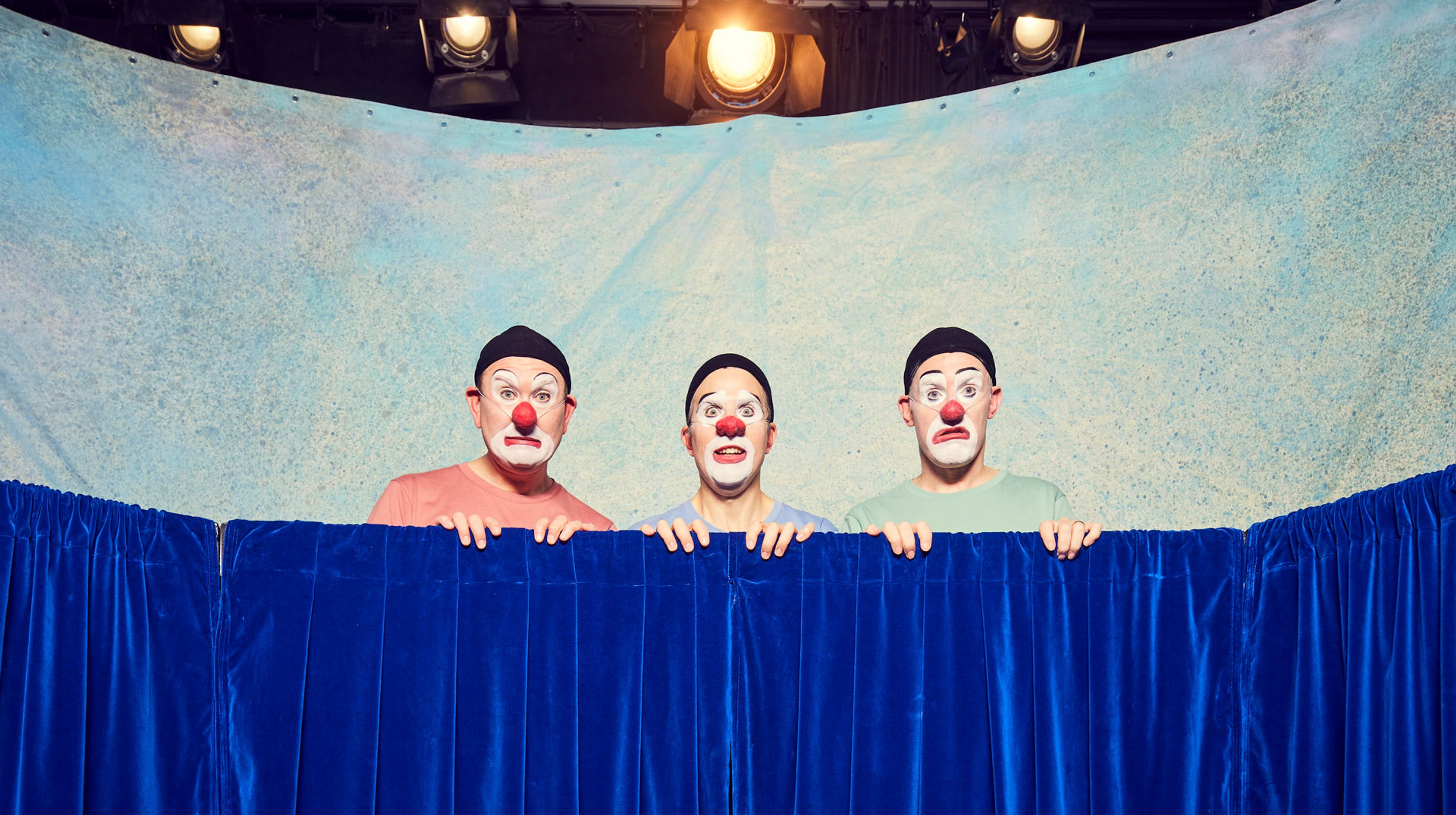 Tre personer i clownsminkning tittar upp över ett skynke. 