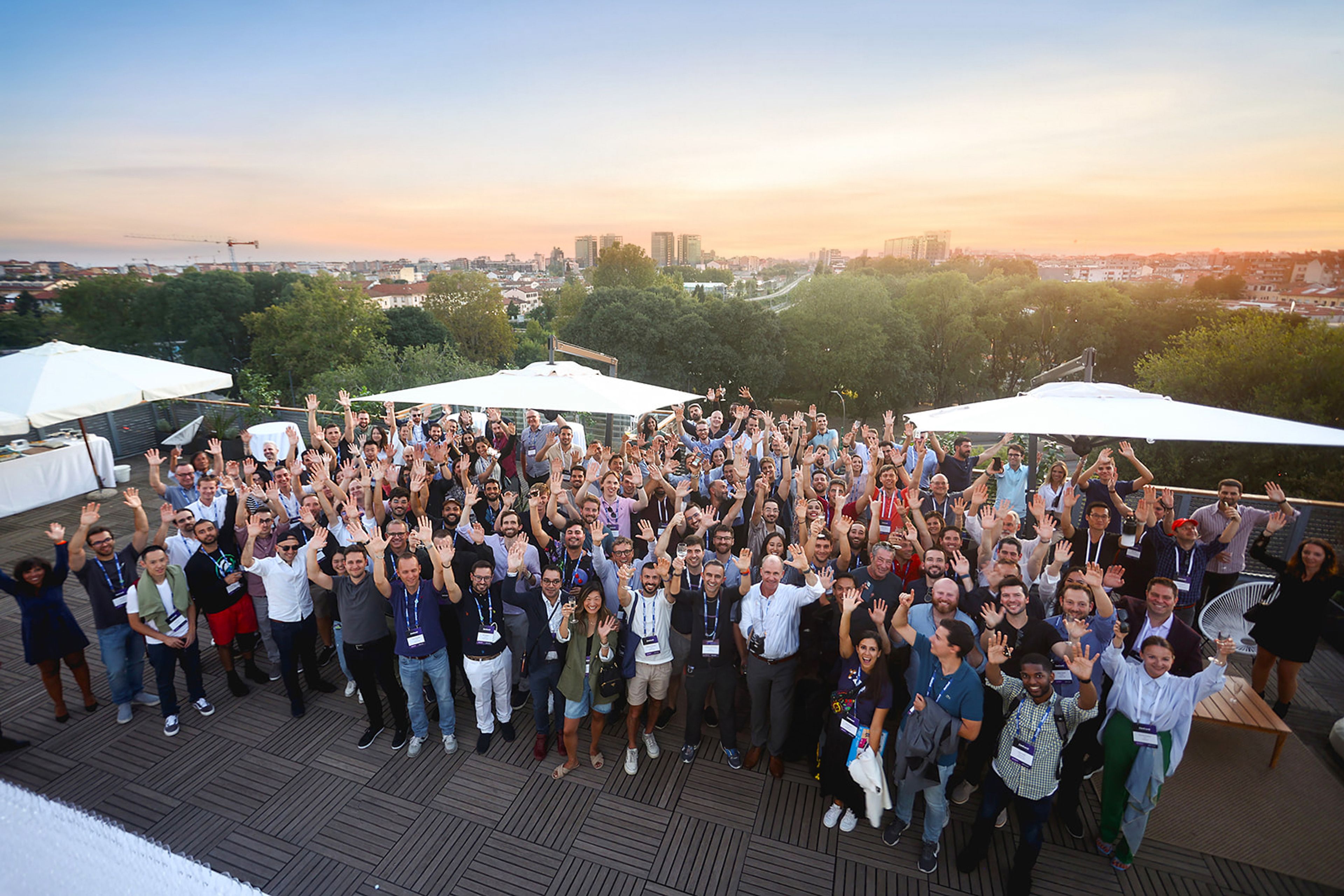 Horizen Labs employees gathered on a rooftop at sunset, smiling and waving during an all-team event, with a city skyline in the background