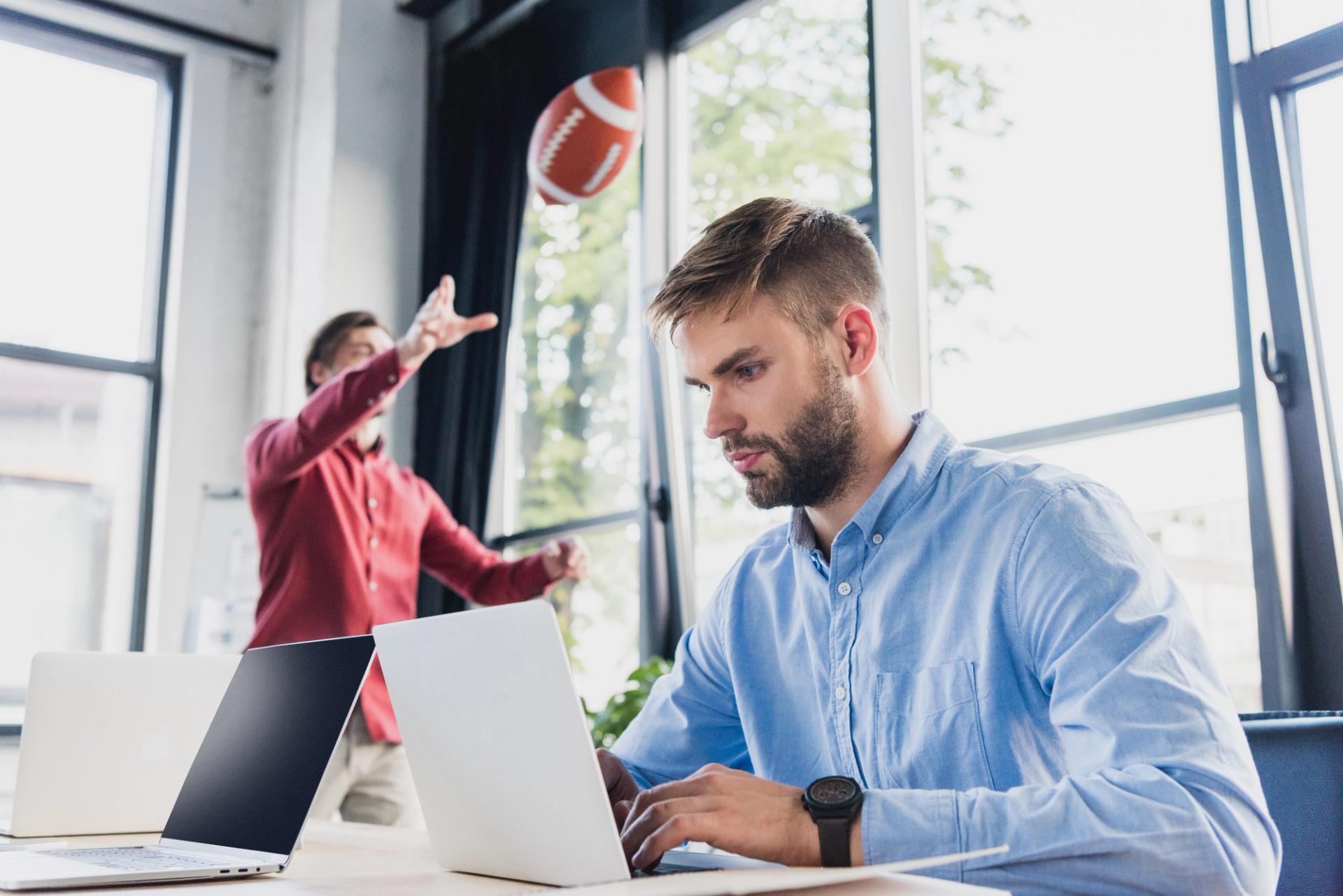 Studente concentrato al laptop durante il corso IFTS Sport Marketing, con un compagno che lancia un pallone da football nello sfondo.