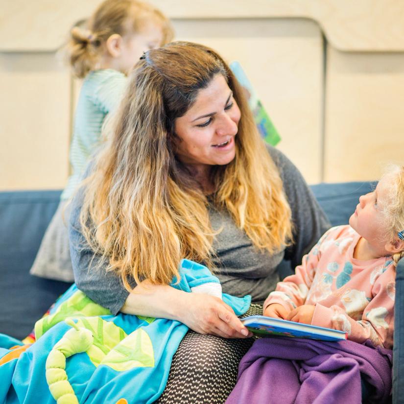 Et barn og en ansatt sitter i en sofa sammen, med en bok i fanget. Foto: Krafttak for sang