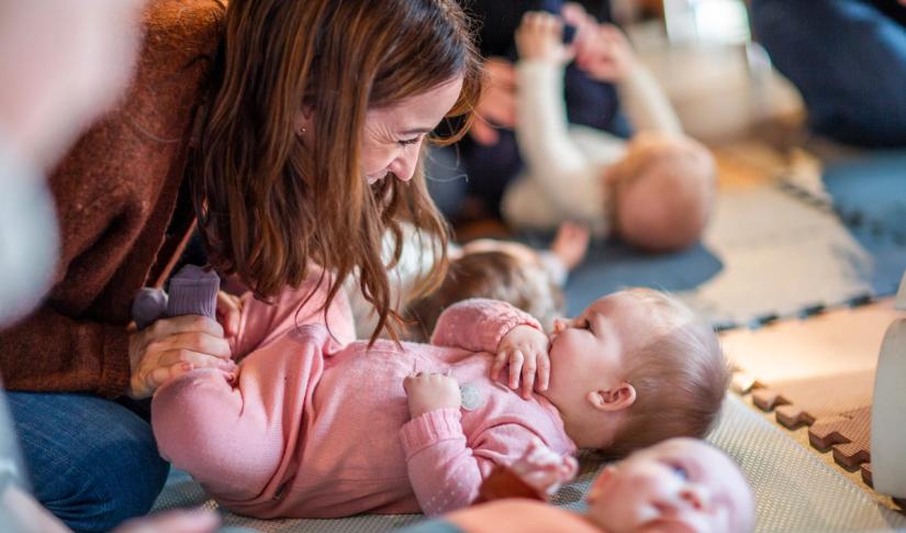 Bilde av babyer som ligger på matter på gulvet og en mor som smiler ned til sitt barn. Foto: Krafttak for sang