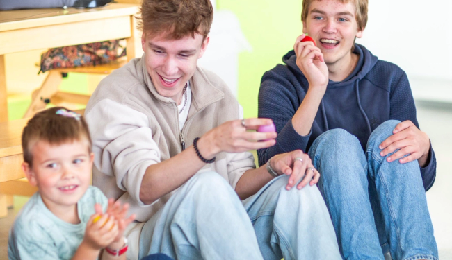 Bilde av to vgs elever som synger og spiller rasleegg med et barn