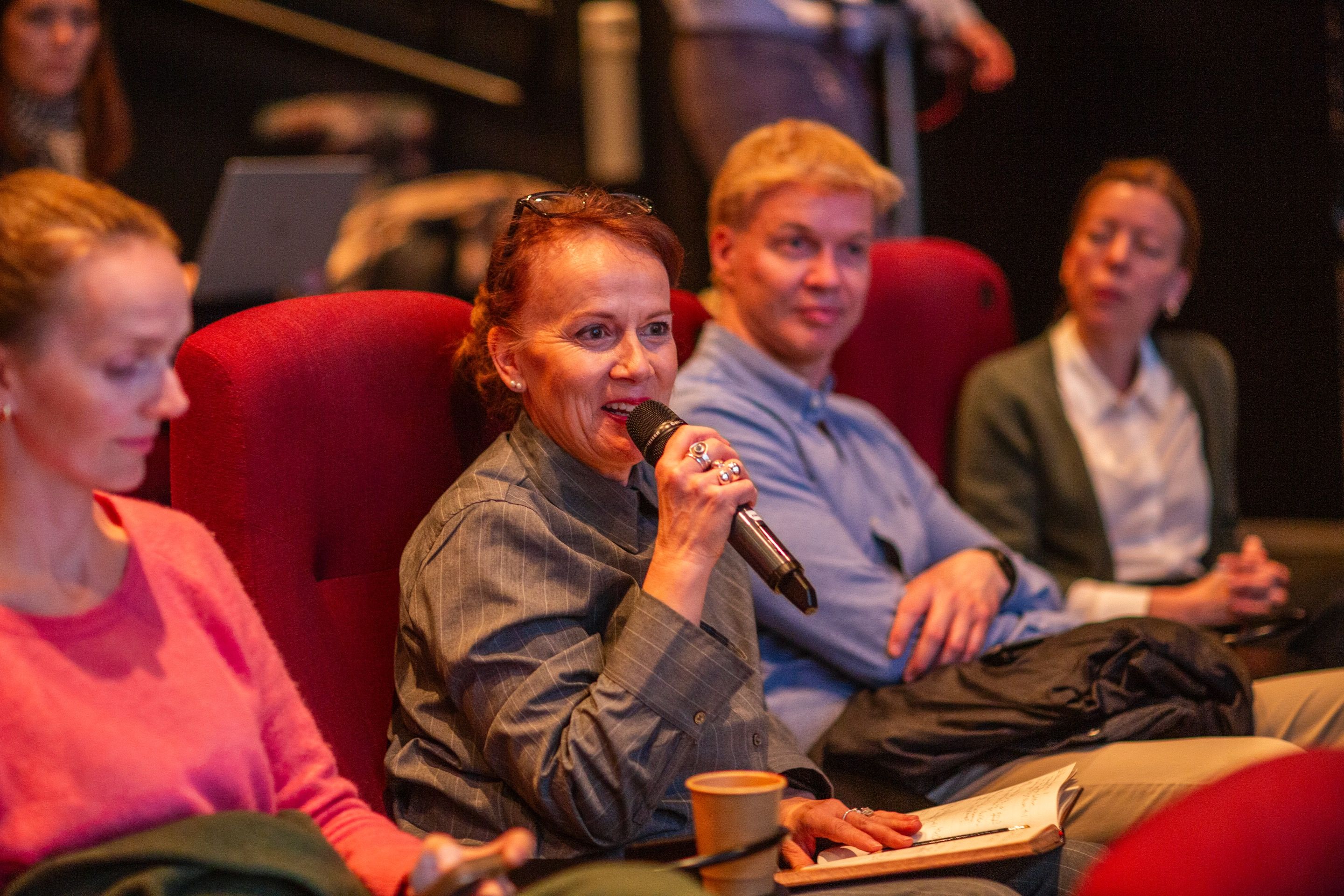 En dame snakker i en mikrofon, det sitter andre voksne rundt henne og lytter. 