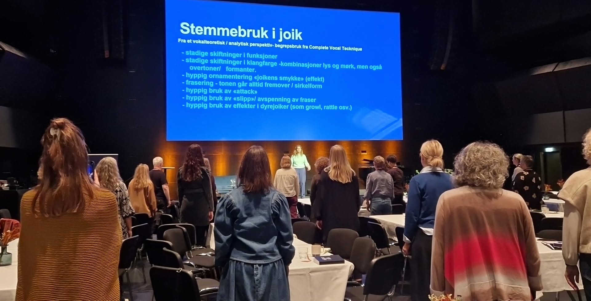 Bilde av en sal der publikum står oppreist. 