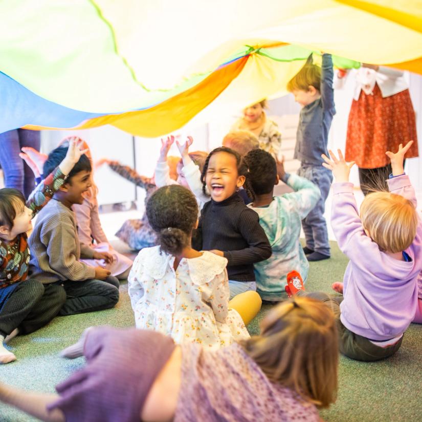 Bilde av en gruppe barn som sitter under en fallskjerm, smiler og ler