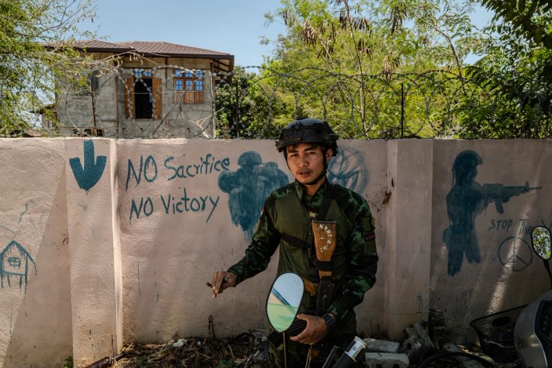 A young KNDF soldier in combat helmet and uniform stands before a concrete wall topped with razor wire, painted with the slogan "No Sacrifice No Victory" and stenciled silhouettes of armed fighters, Karenni State, Myanmar, 2024.