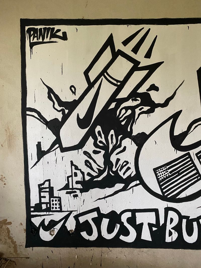 Nike's swoosh redrawn as ordnance. A cityscape below, a US flag to the right, and a familiar slogan rewritten at the bottom. PANIK signs his name at the top. The wall does the rest.