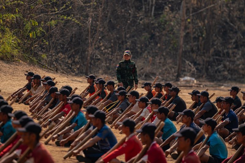 A female Karenni Nationalities Defense Force officer in camouflage uniform stands among rows of seated male recruits holding wooden batons during an outdoor training drill in Myanmar, March 2024.