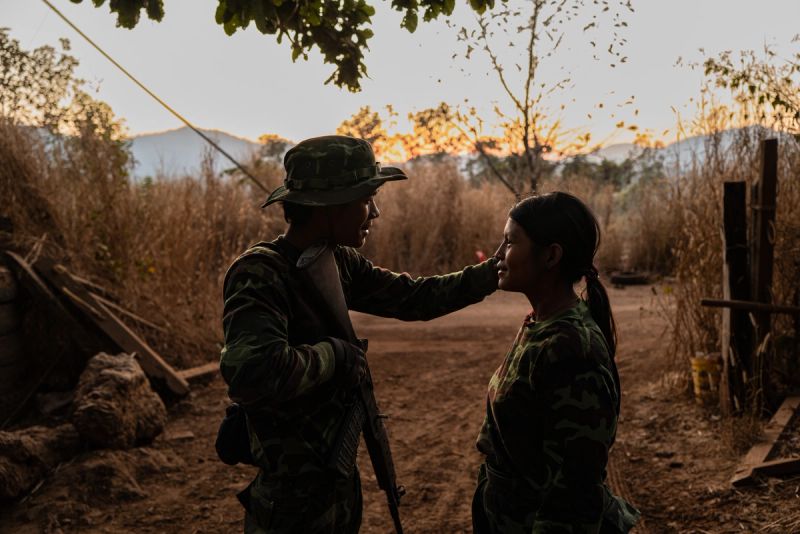 Two KNDF fighters in camouflage uniforms share a tender moment at sunset in a rural outpost in Karenni State, Myanmar. One fighter gently touches the face of the other against a backdrop of dry grass and mountains, 2023.