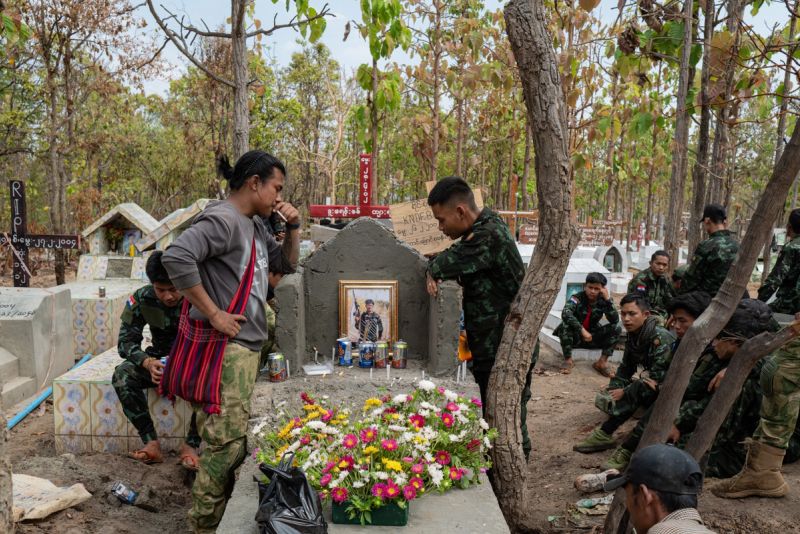 KNDF soldiers gather around a freshly constructed grave adorned with flowers, candles, beer cans, and a framed portrait of the fallen fighter in a woodland cemetery in Myanmar, 2024.