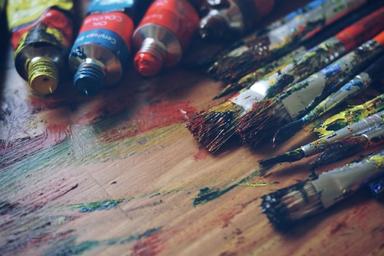 Image of many messy and colourful paint covered paintbrushes and tubes of paint lying on a table.