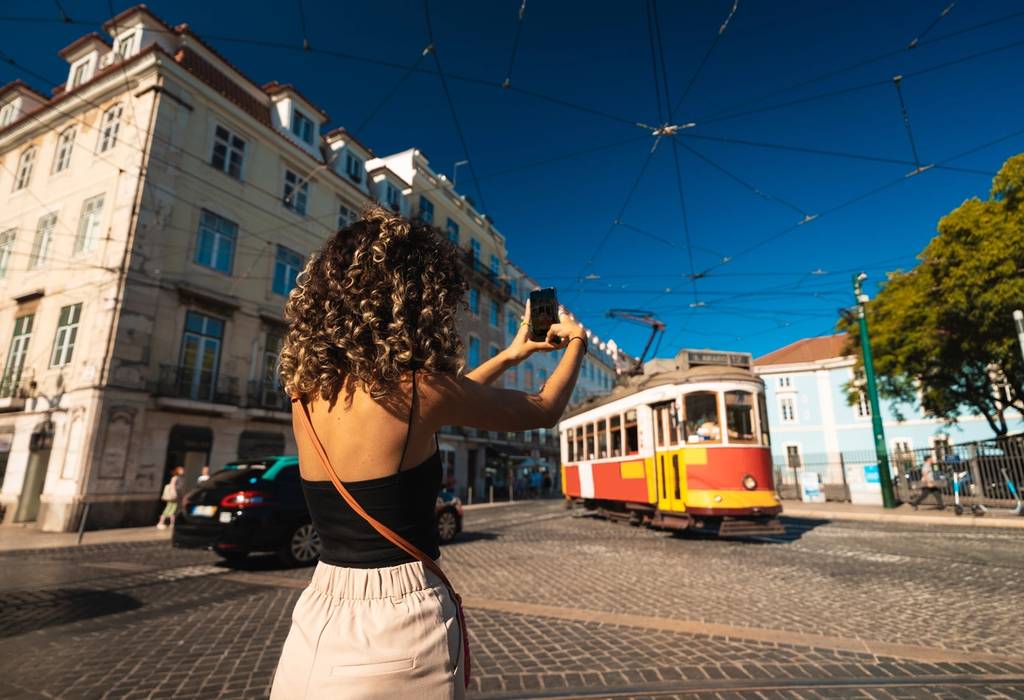A picture of a tourist taking a photo of the iconic yellow tram in Lisbon, Portugal whilst a taxi drives past