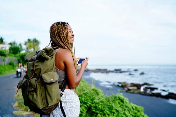 A cheerful female backpacker holding smartphone in her hands while trekking along a black-sand beach