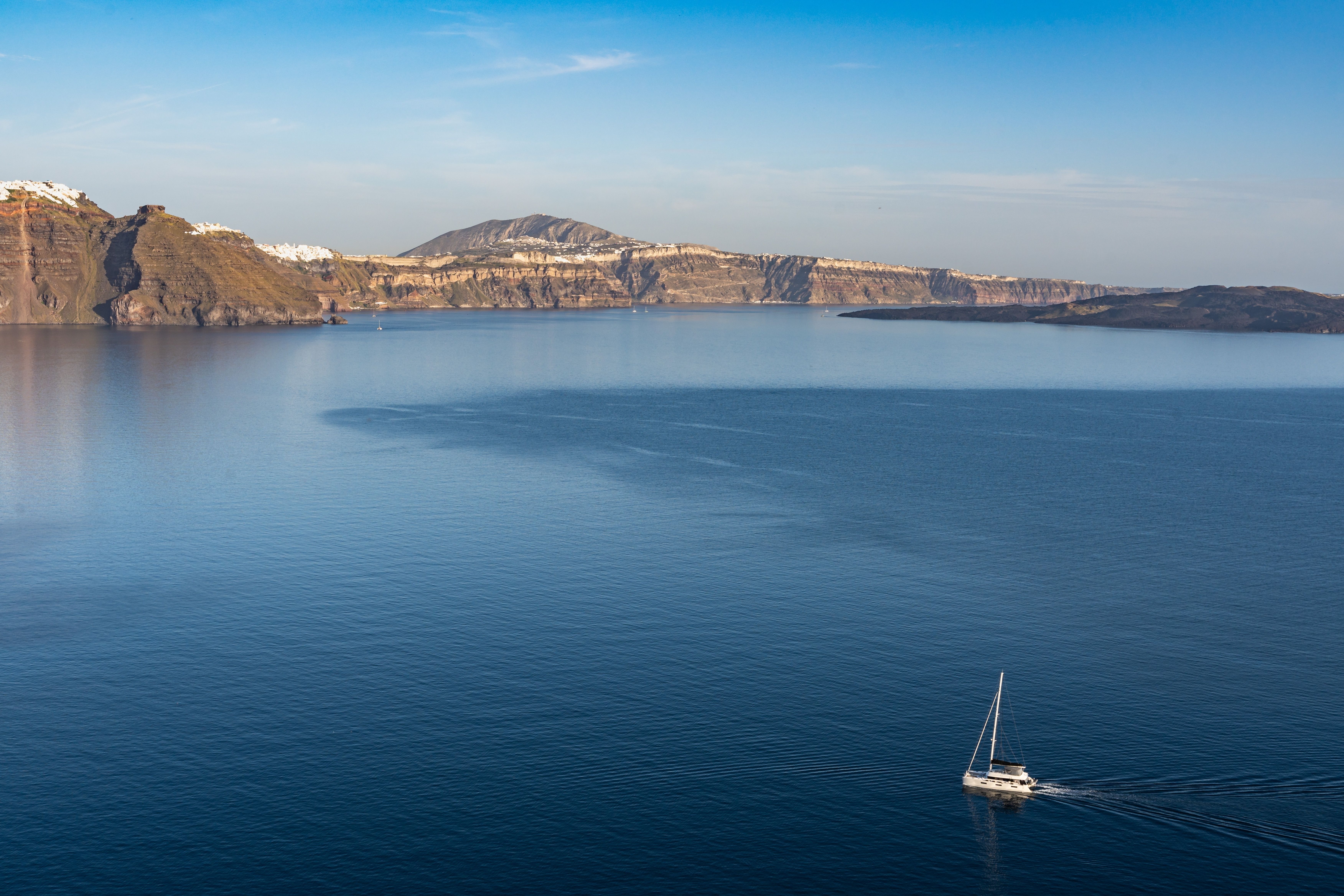 A catamaran sails across the deep blue waters towards the rocky coast of Santorini