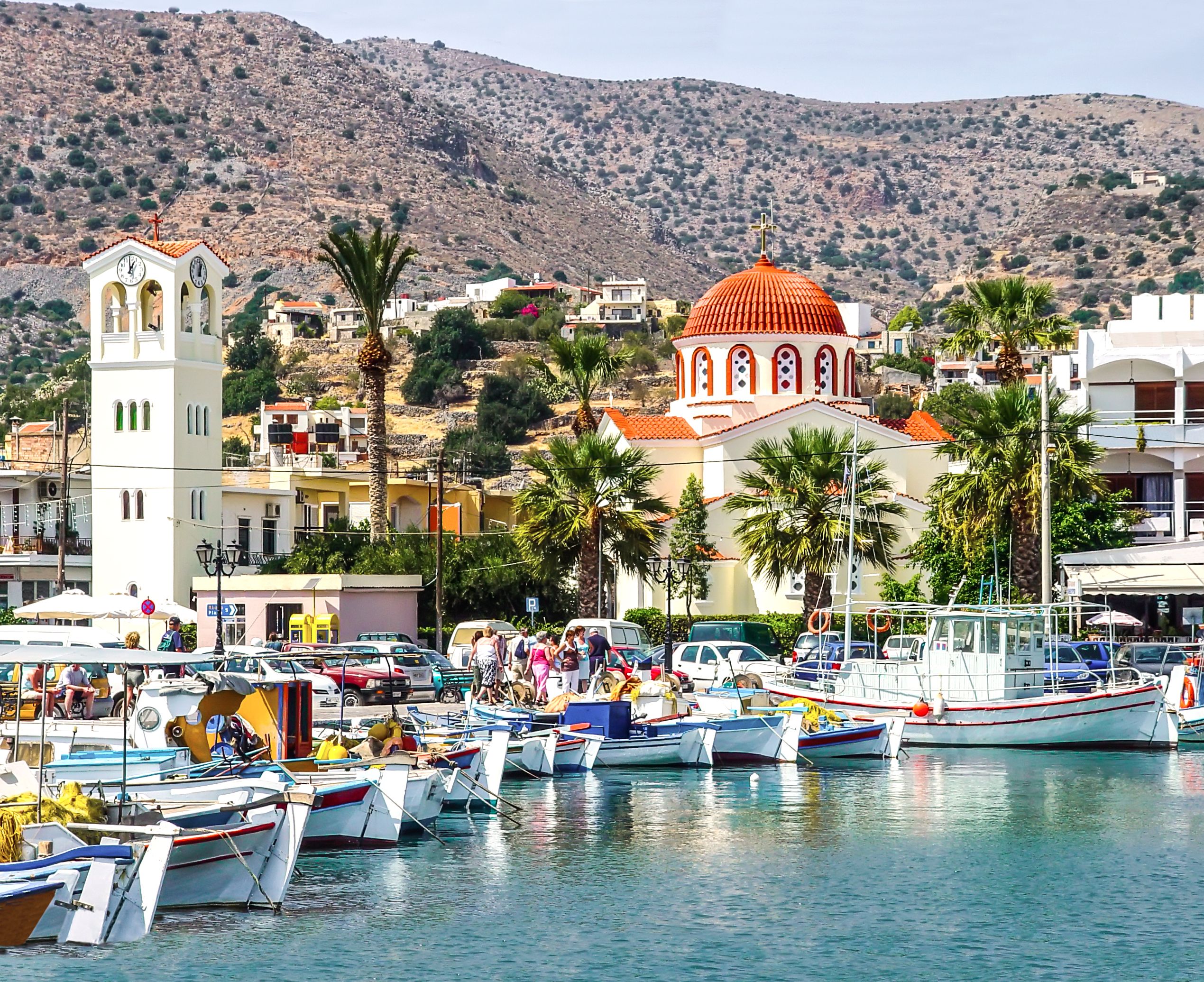 View of boats bobbing in the marina of a Greek island town with a bell tower and red-domed church in the background