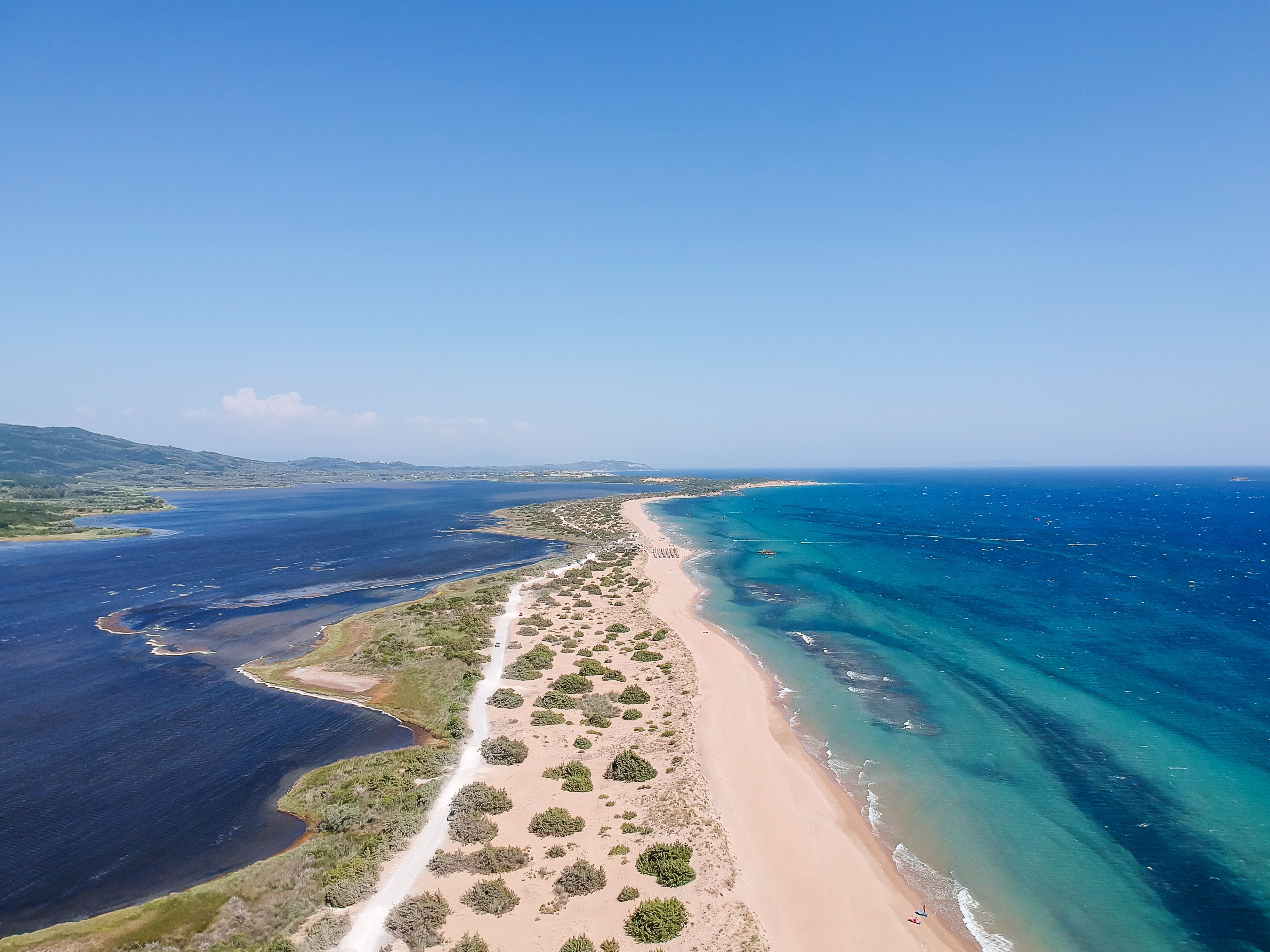 Drone view of a narrow shrub-dotted sandbar separating a deep blue lagoon and the clear, turquoise Med.