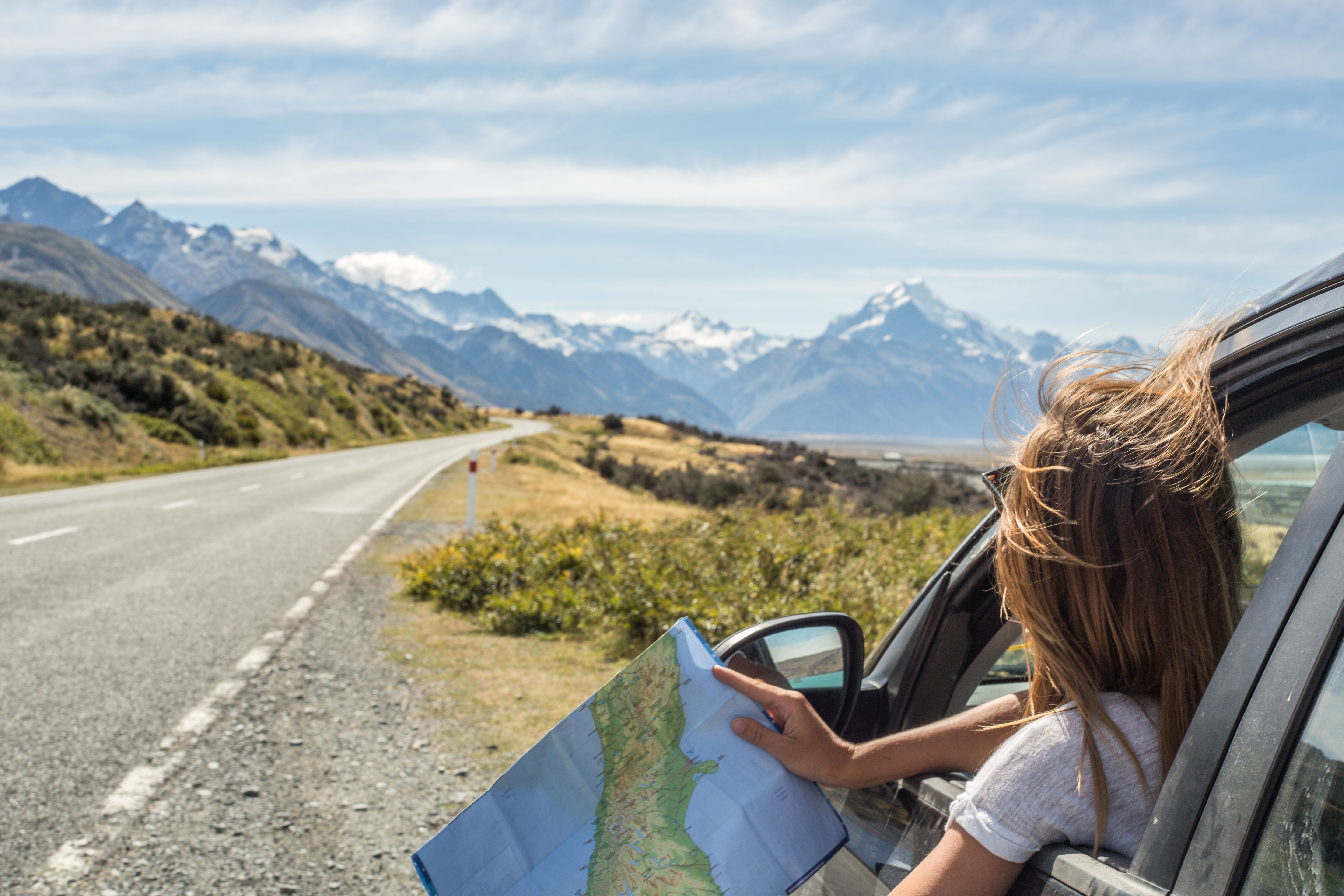 A woman pulled over by the side of a road to look at a paper map with snow-capped mountains in the distance