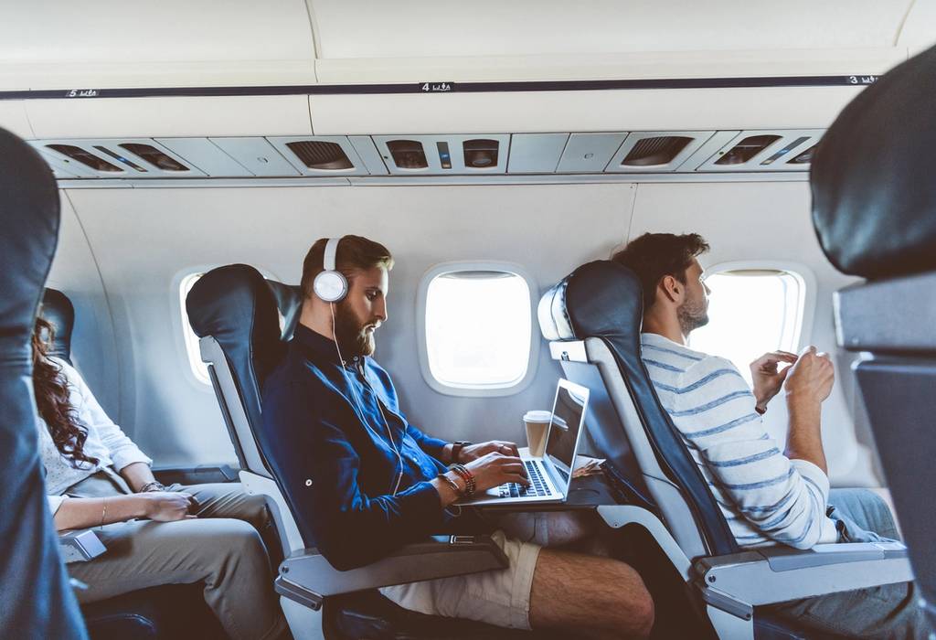 A male passenger using his laptop wearing headphones during a flight with other people sat around him