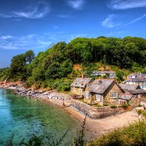 A view of a picturesque seaside village in Cornwall surrounded by green trees with a small beach lapped by clear blue water