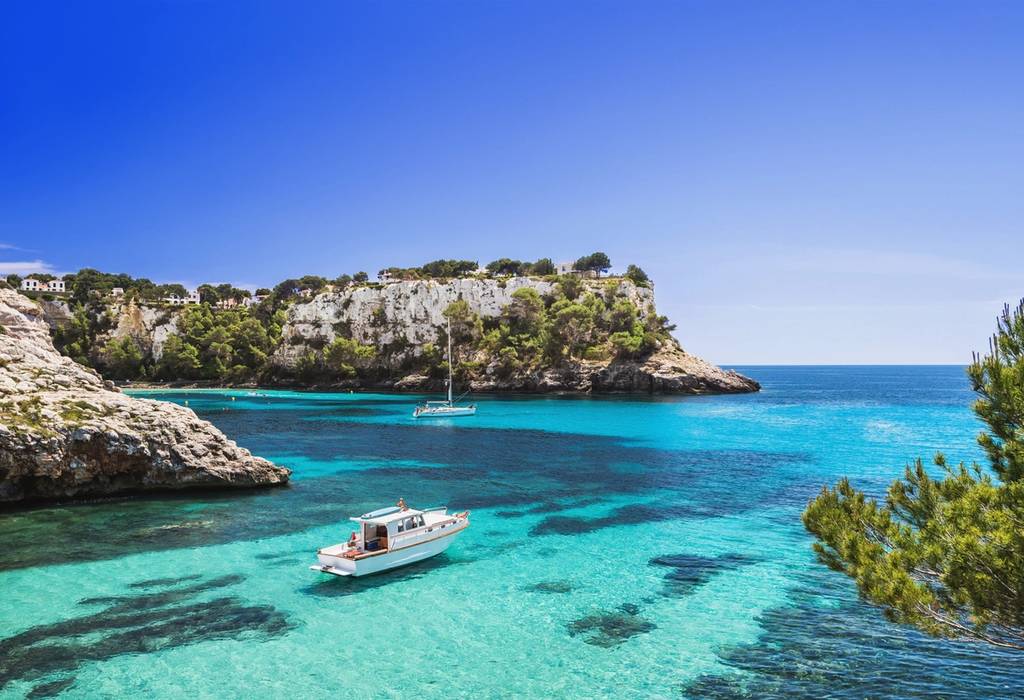View of two boats in the clear, shallow waters of Cala Galdana beach on a sunny day in Menorca