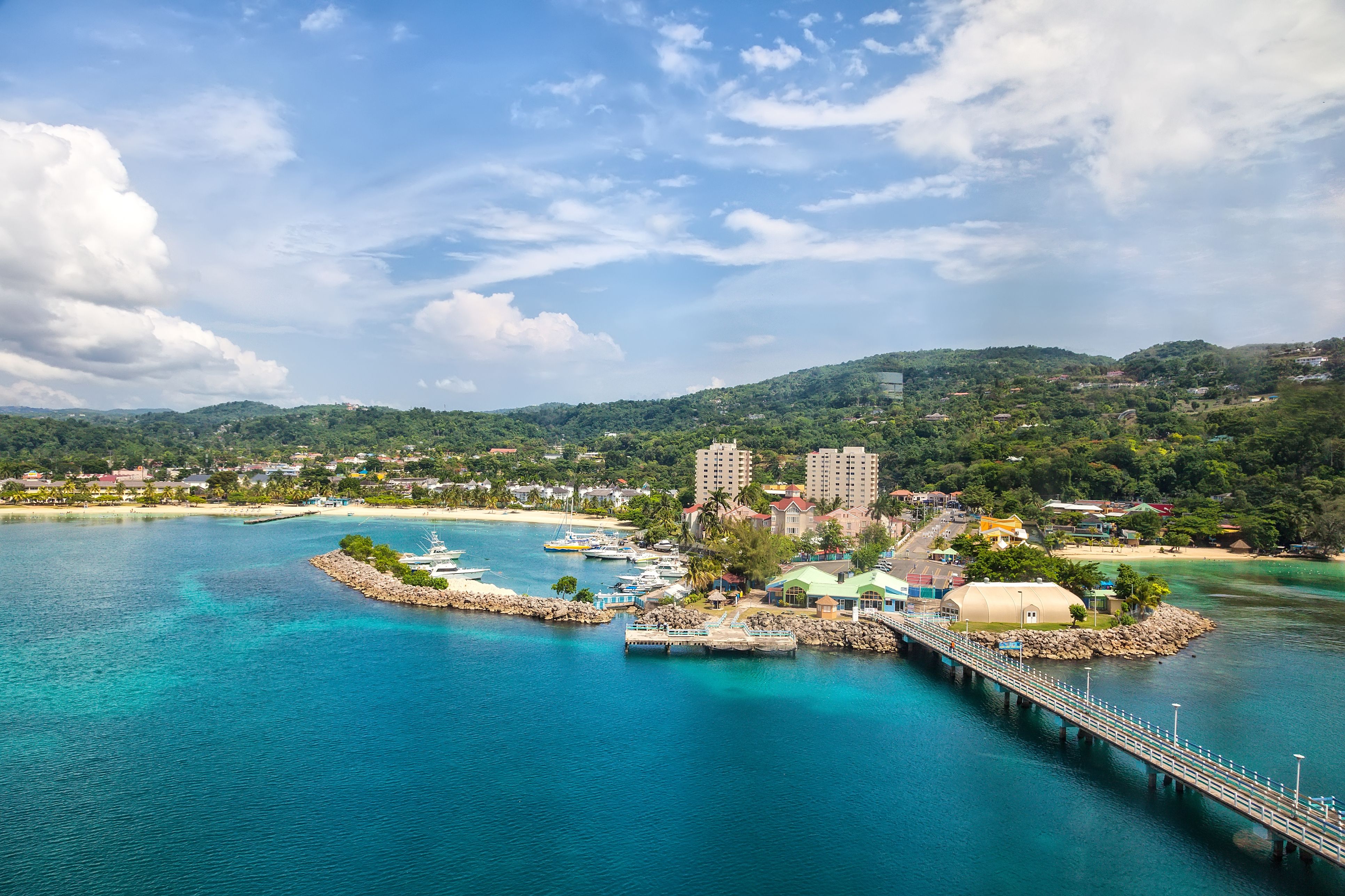 Aerial view of a coastal town in Jamaica with a cruise port, over-water bridge and a long sandy beach, all on a backdrop of tree-filled hills