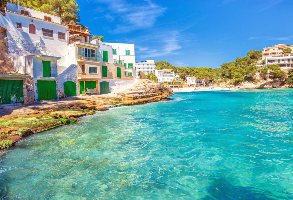 A view of traditional white-washed boathouses, apartments and beach at Playa Santanyi in Majorca with azure-coloured sea and clear blue sky