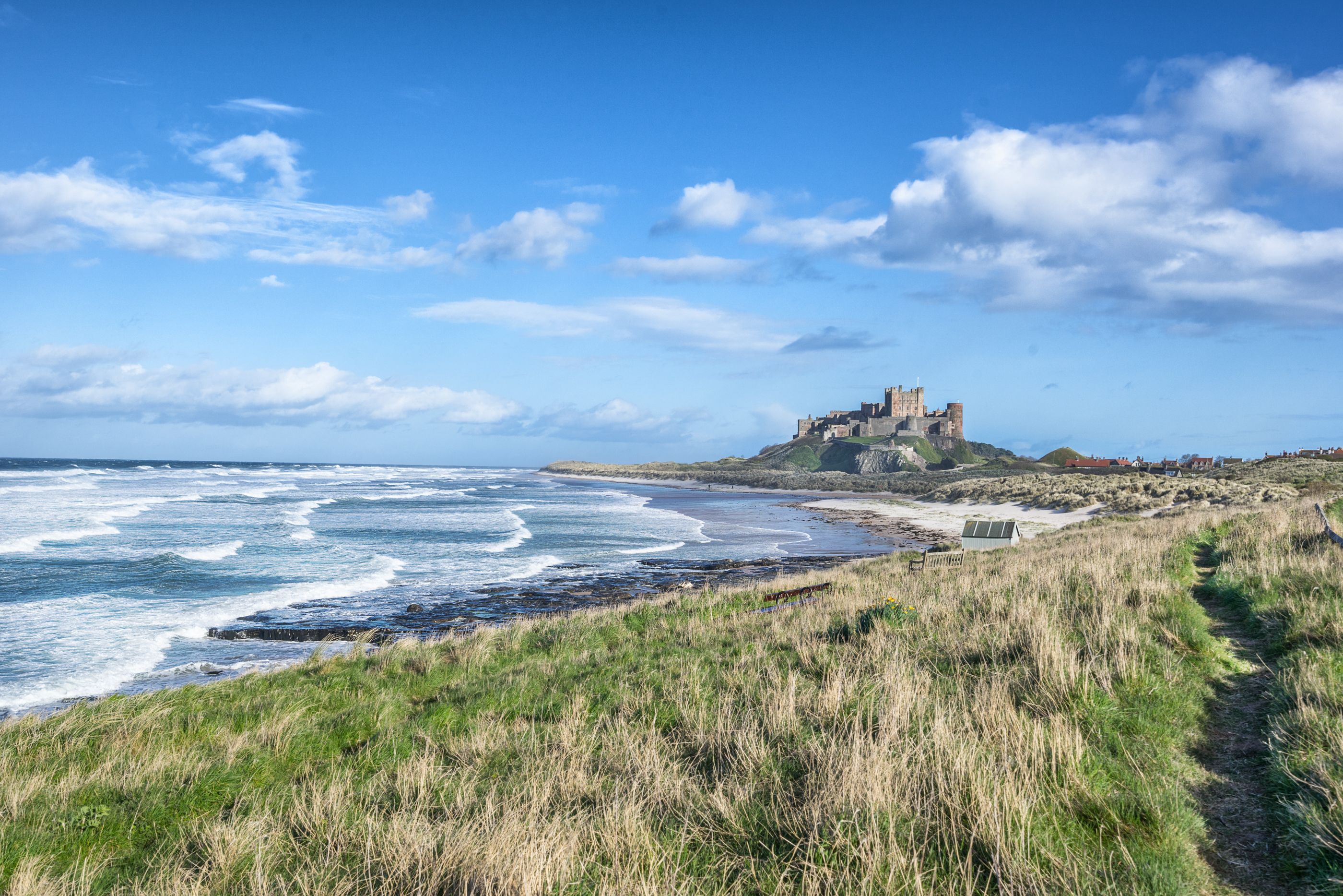 View across grassy cliffs towards a pristine beach with an imposing castle overlooking it all
