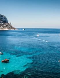 View of yachts sailing in the Mediterranean off the shore of Cala d'Hort beach in Ibiza with the rocky Es Vedra island in the background