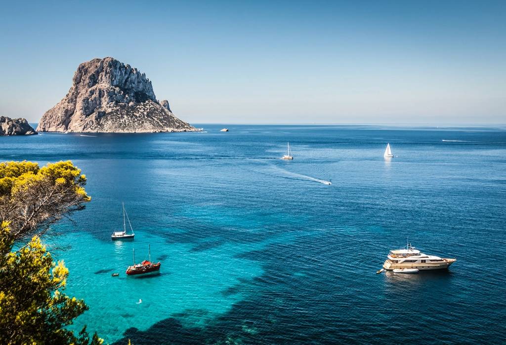 View of yachts sailing in the Mediterranean off the shore of Cala d'Hort beach in Ibiza with the rocky Es Vedra island in the background