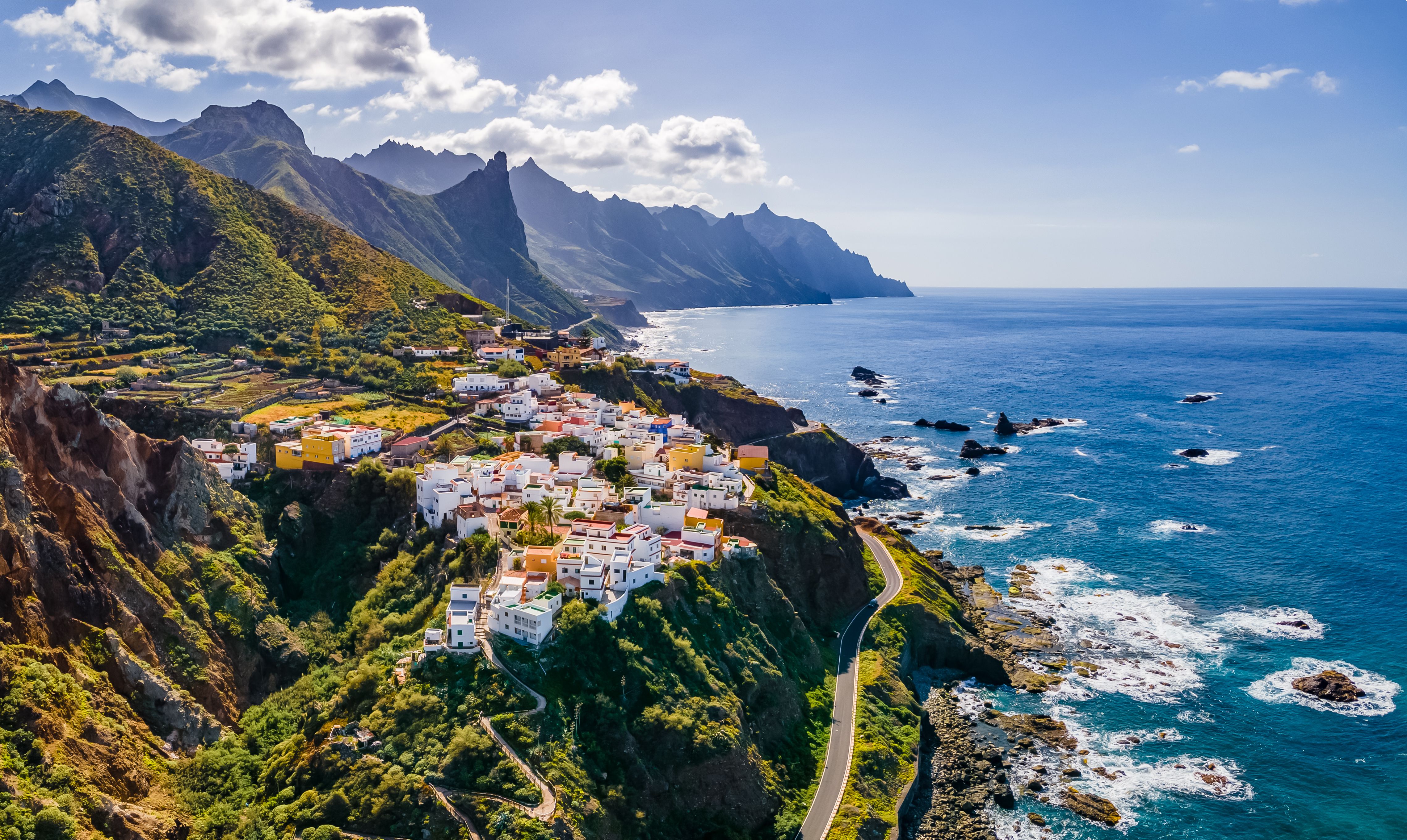 Aerial drone view of a clifftop hamlet surrounded by sharp mountain peaks and the powerful Atlantic Ocean.
