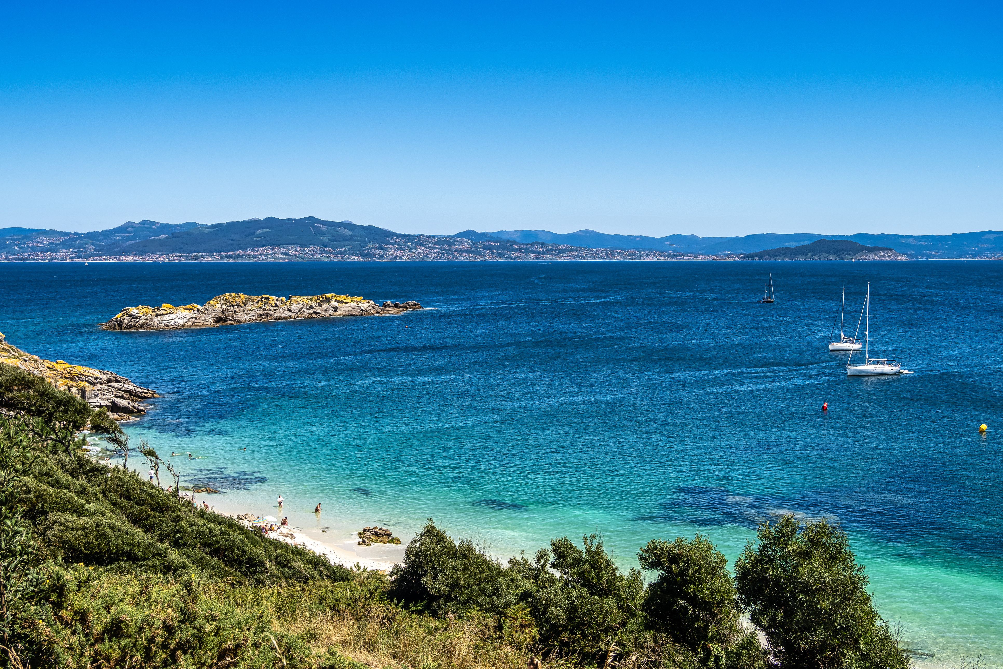 View across a calm bay with yachts bobbing in the distance and a few people on a white-sand beach.