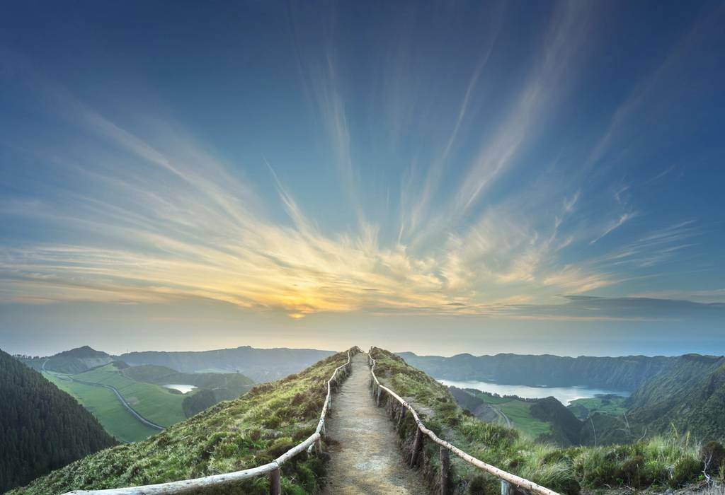 A hiking trail stretches into the distance at a viewpoint across a crater lake in the Azores as the sun rises