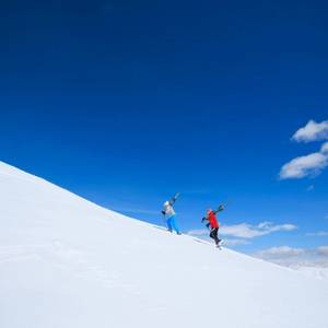 A view of two people carrying their skies as they walk up the side of a snowy mountain on a sunny winter day in Livigno ski resort of the Italian Alps