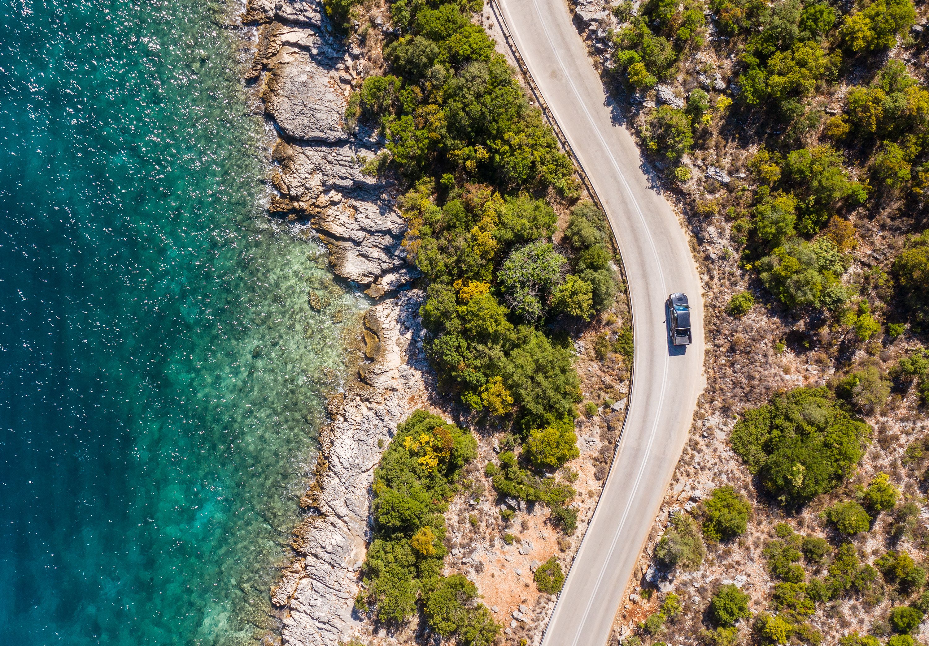 A bird's eye view of a pick-up truck driving along a coastal road in Kefalonia, Greece with calm blue sea