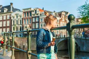 A young girl holding an ice-cream and looking over a canal in Amsterdam with traditional Dutch buildings out of focus in the background