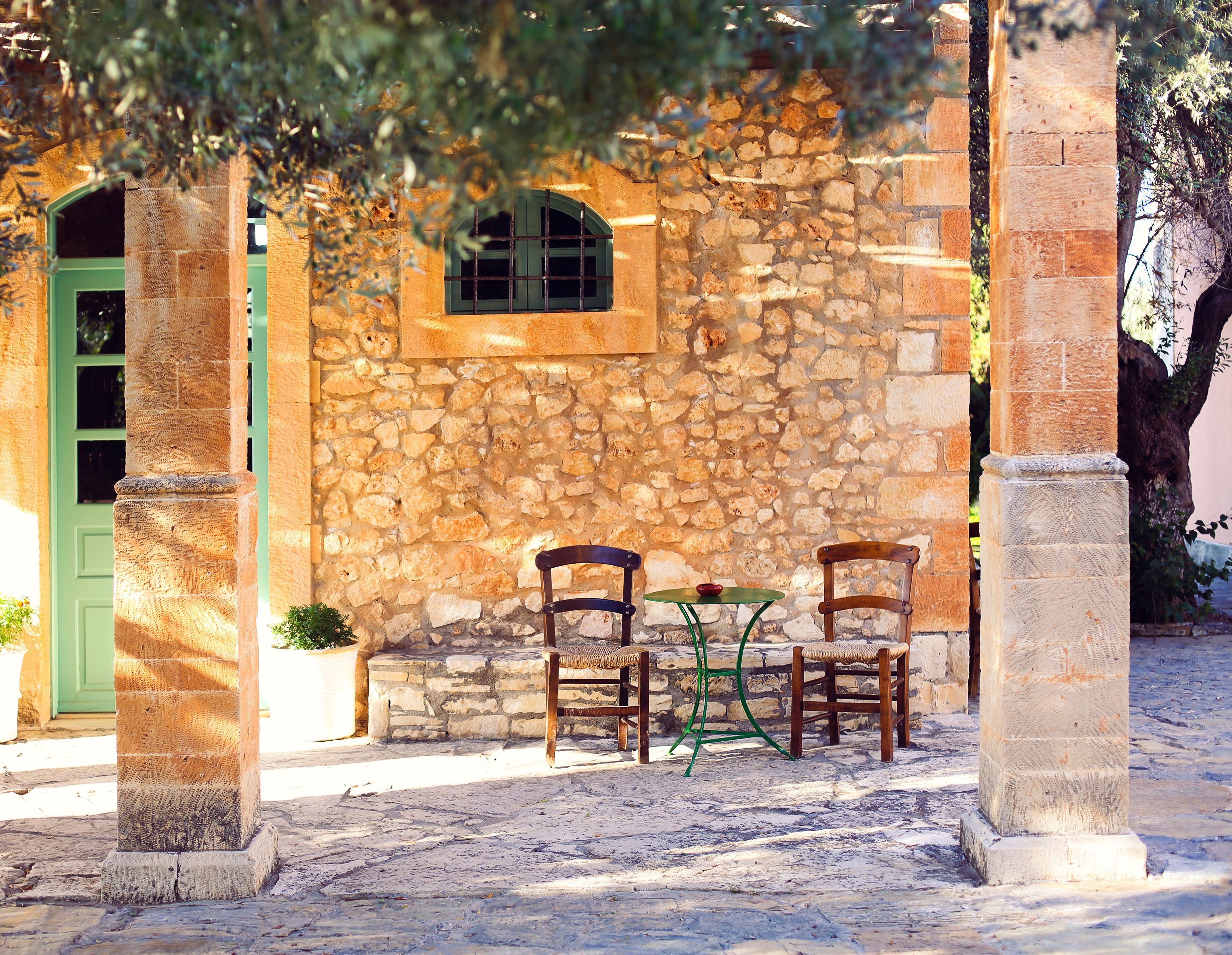 View of two wooden chairs and a small green table in front of a honey-coloured stone taverna in Crete.