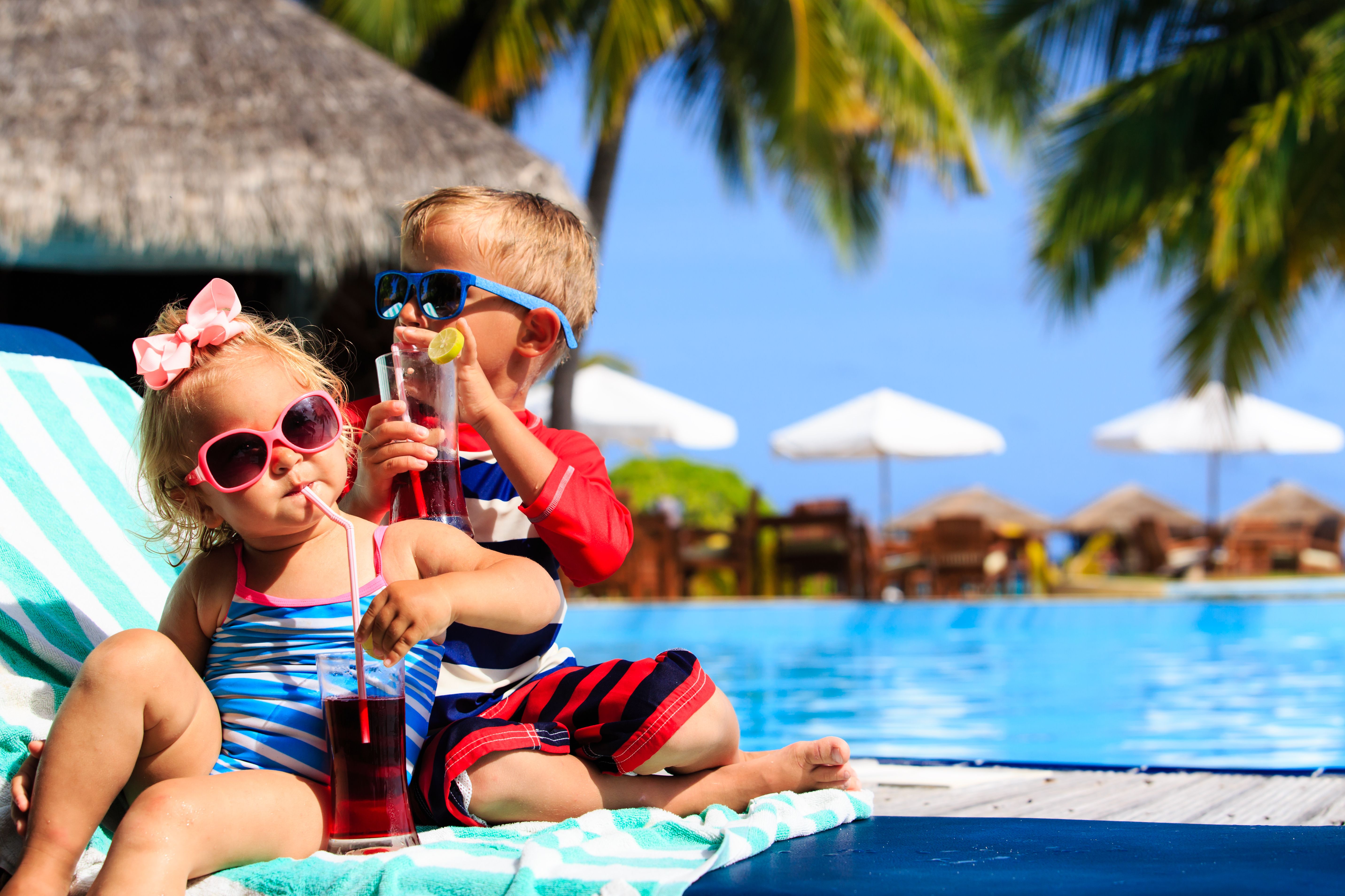 Two young children relax by a pool in a tropical beach resort with drinks