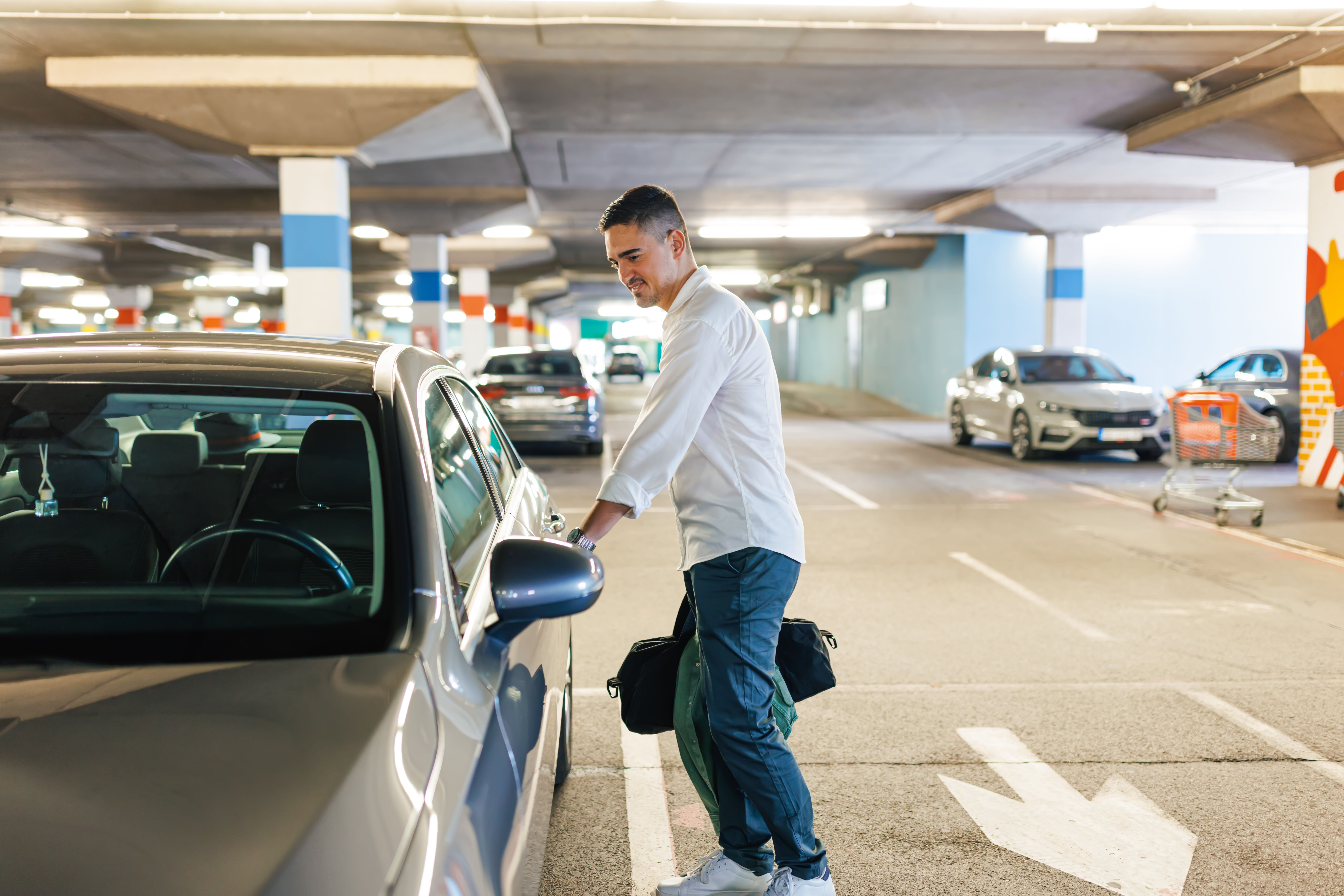 A young man stands in an underground garage with a bag in hand, opening his car door and smiling.