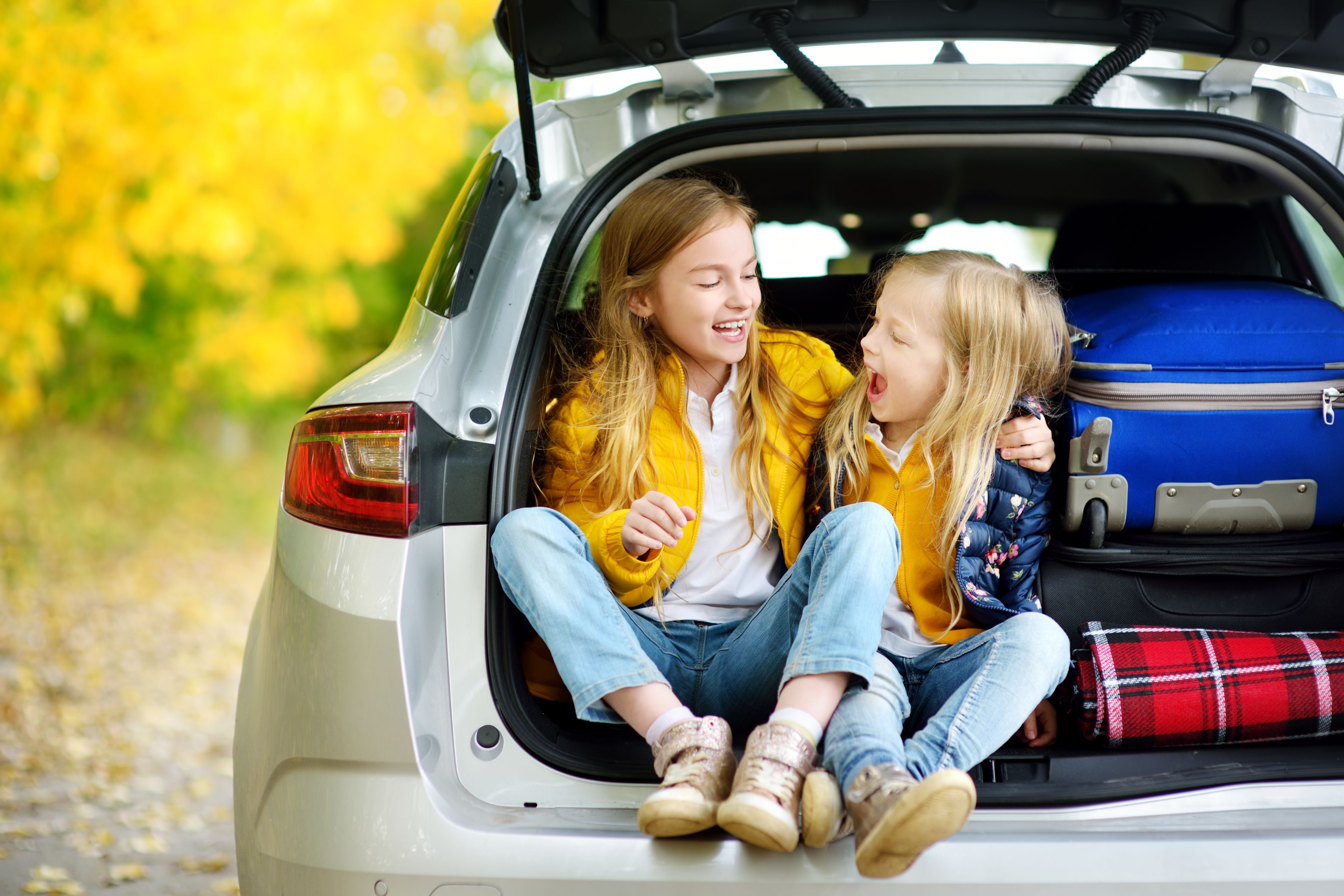 Two girls laughing in the back of an open car trunk next to luggage on a family road trip holiday