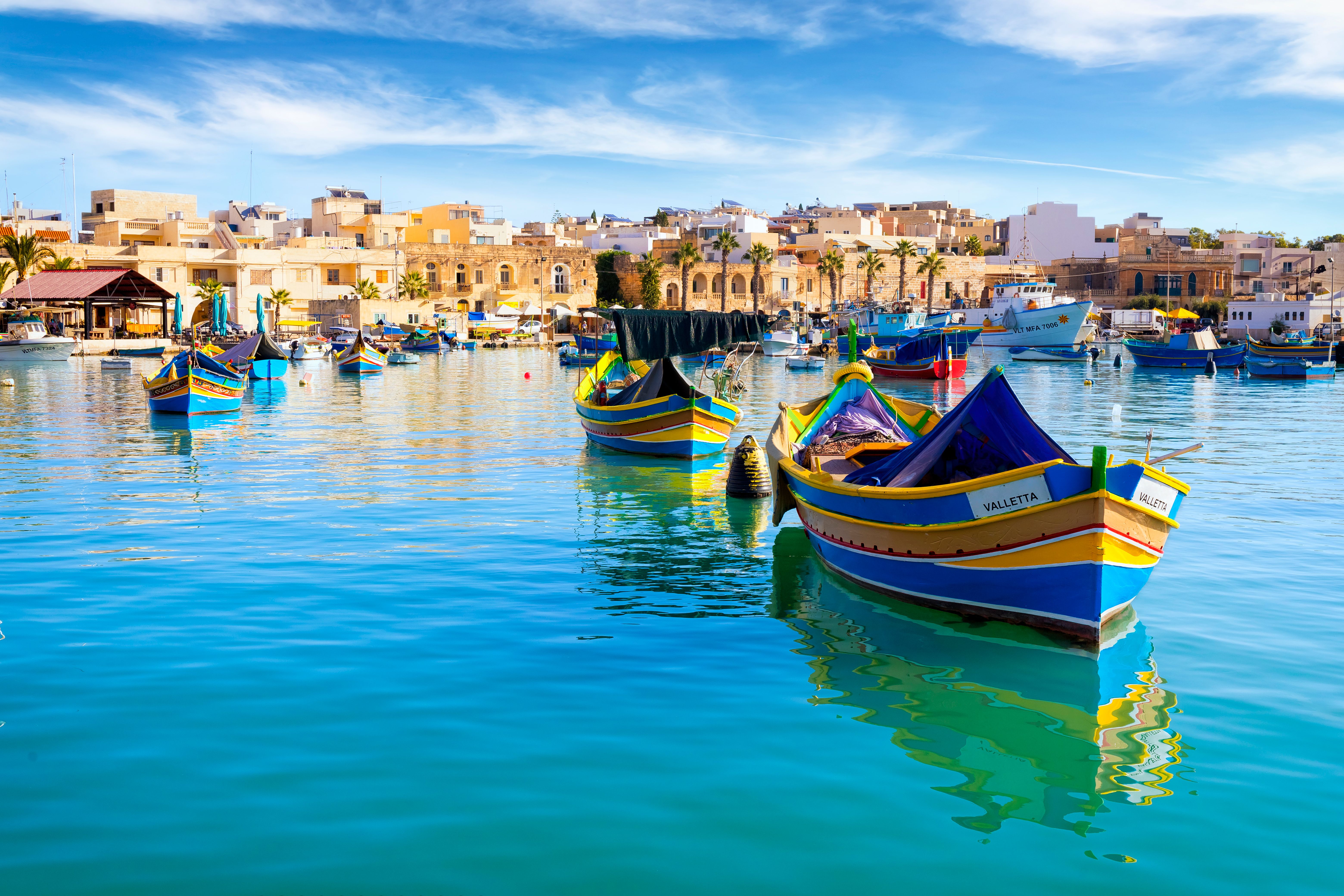 A view looking across the water of Marsaxlokk Fishing Village in Malta with colourful traditional fishing boats moored in the harbour