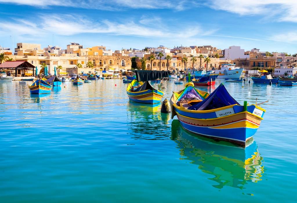 A view looking across the water of Marsaxlokk Fishing Village in Malta with colourful traditional fishing boats moored in the harbour