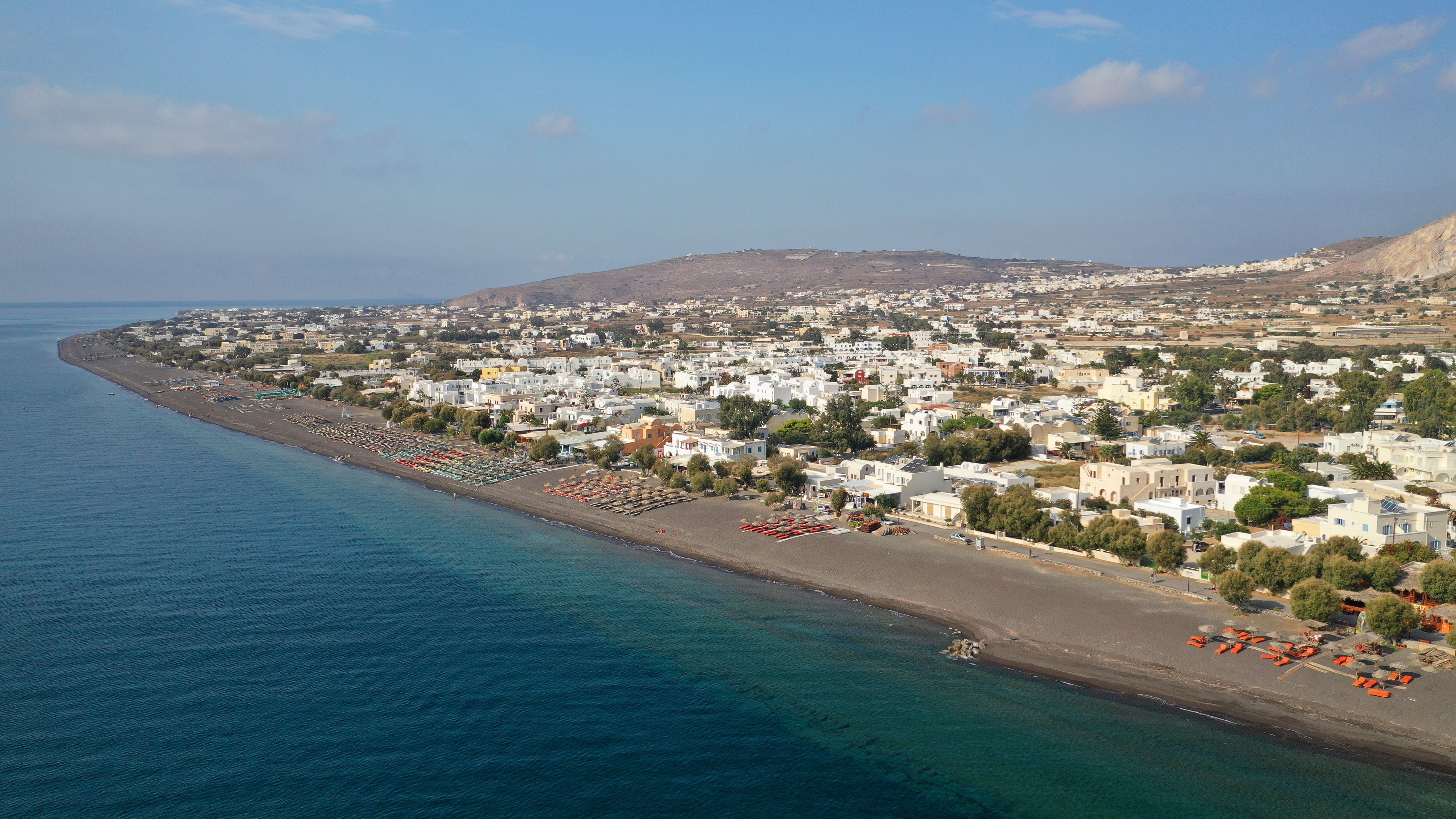 Aerial view of a sunbed-lined black-sand beach backed by a low-rise village of whitewashed village in Santorini