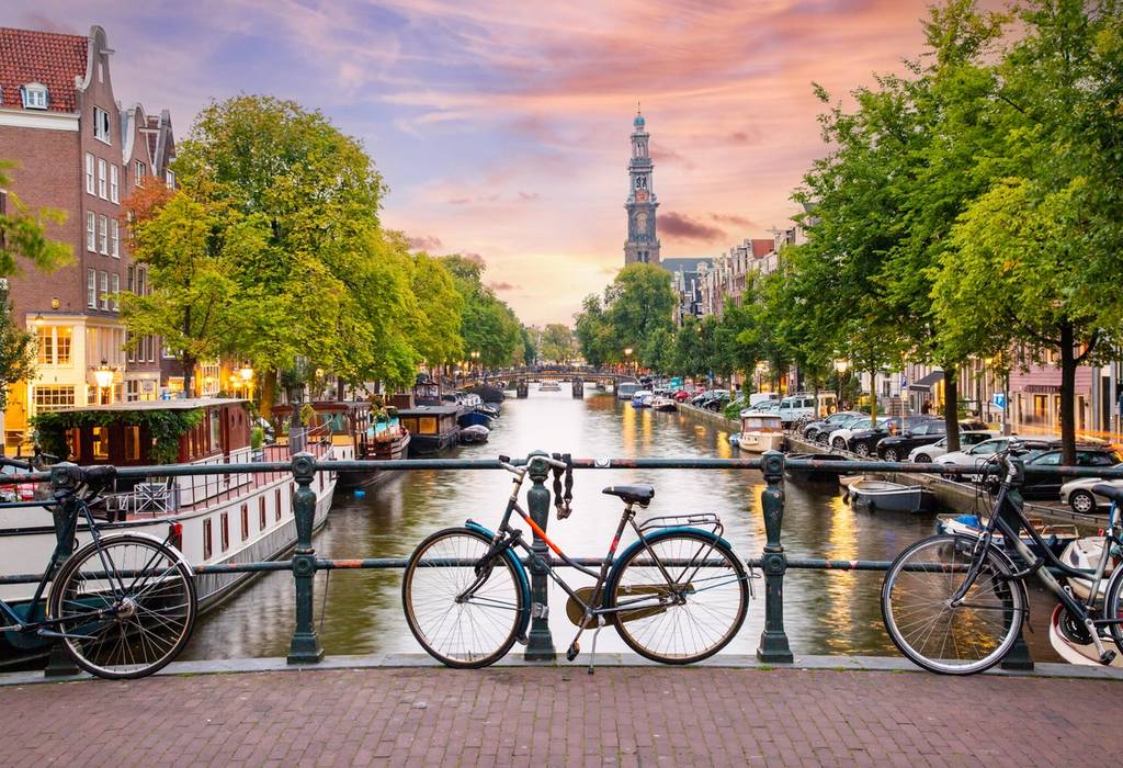 Sunset in Amsterdam with Bicycles parked on a bridge over a canal in the city centre, with typical Dutch houses on the background and streets illuminated with lights