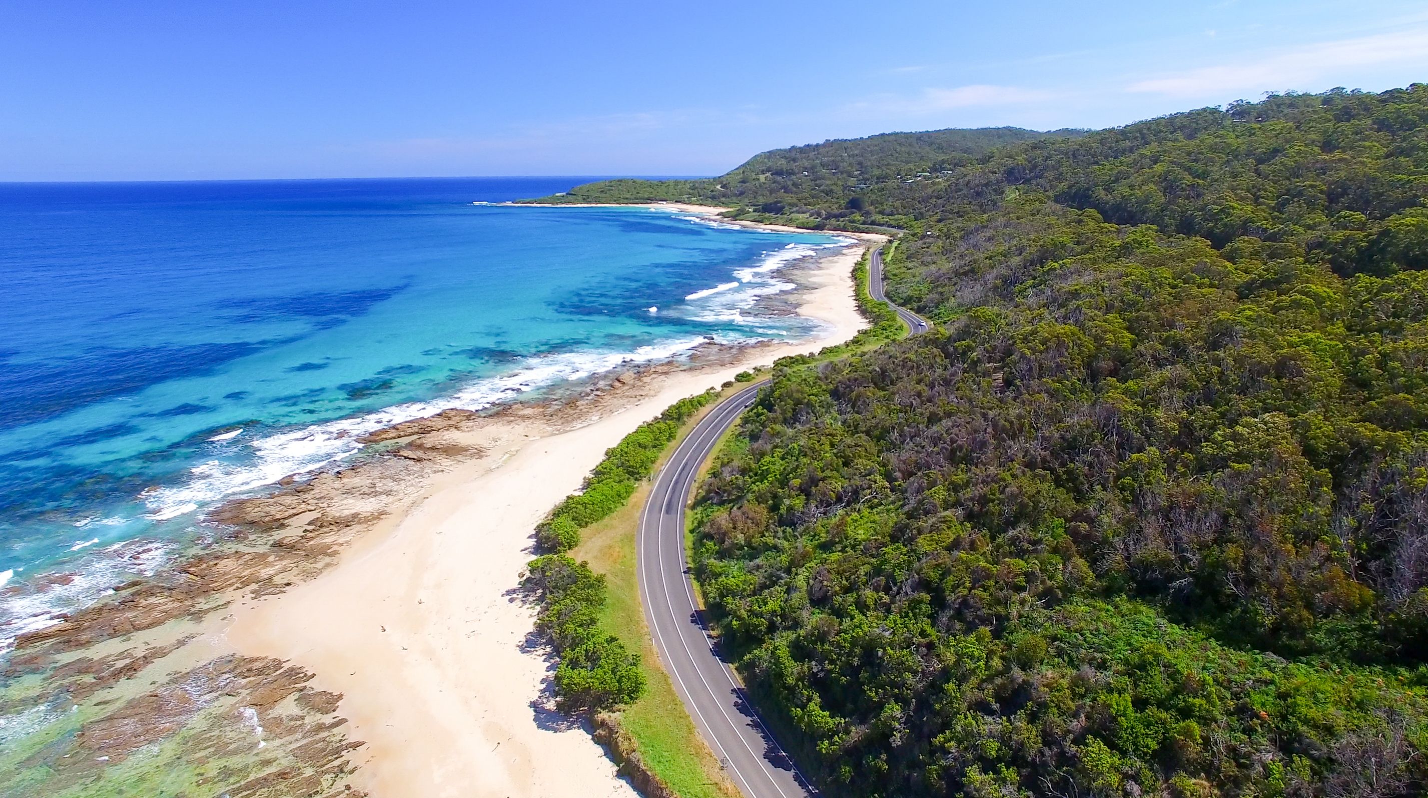 Aerial view of a winding road that hugs the south coast of Australia with bush on the right side and a golden beach to the left