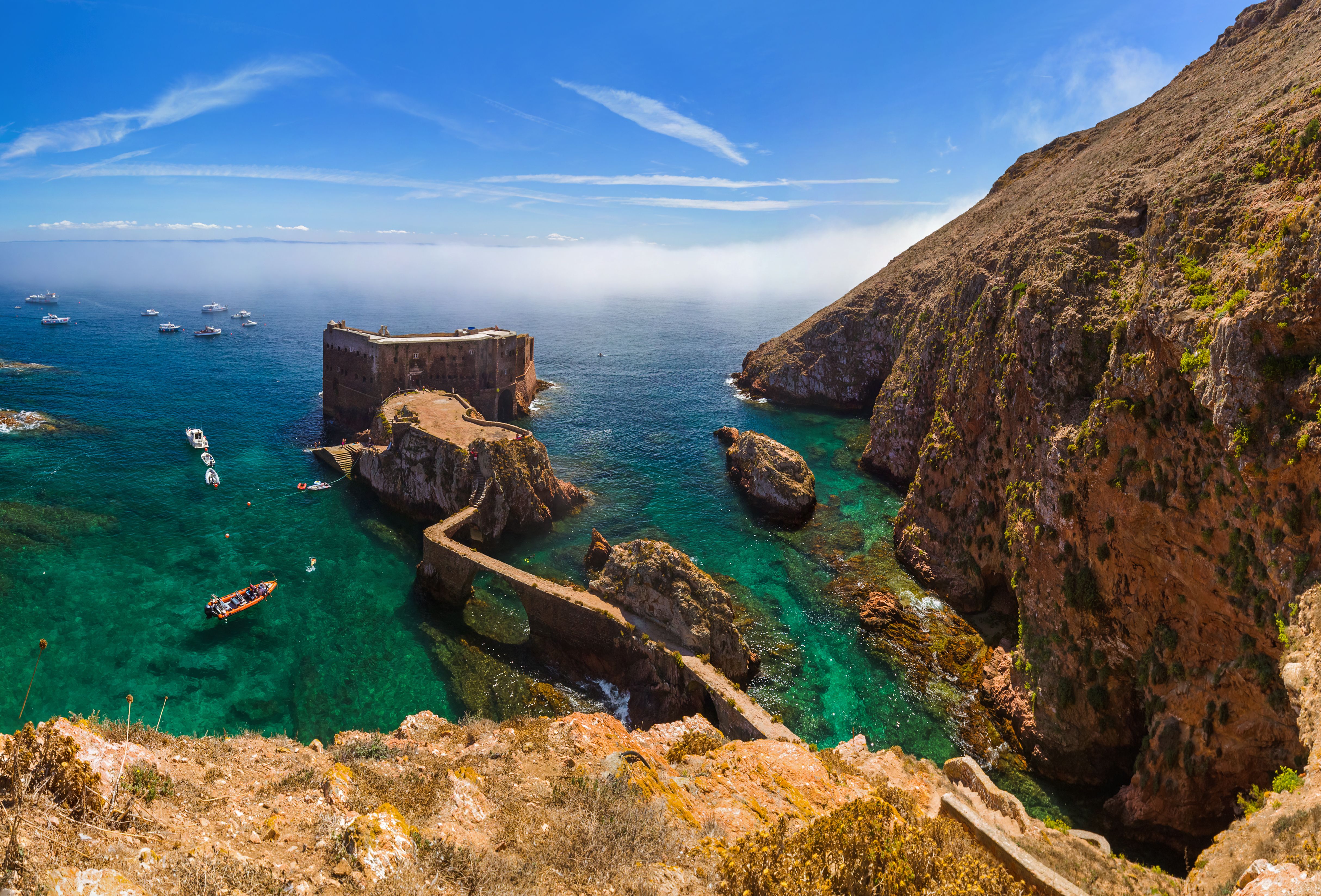 View looking down from a cliff towards a fort in the Atlantic Ocean that's connected to the mainland by a narrow walkway.