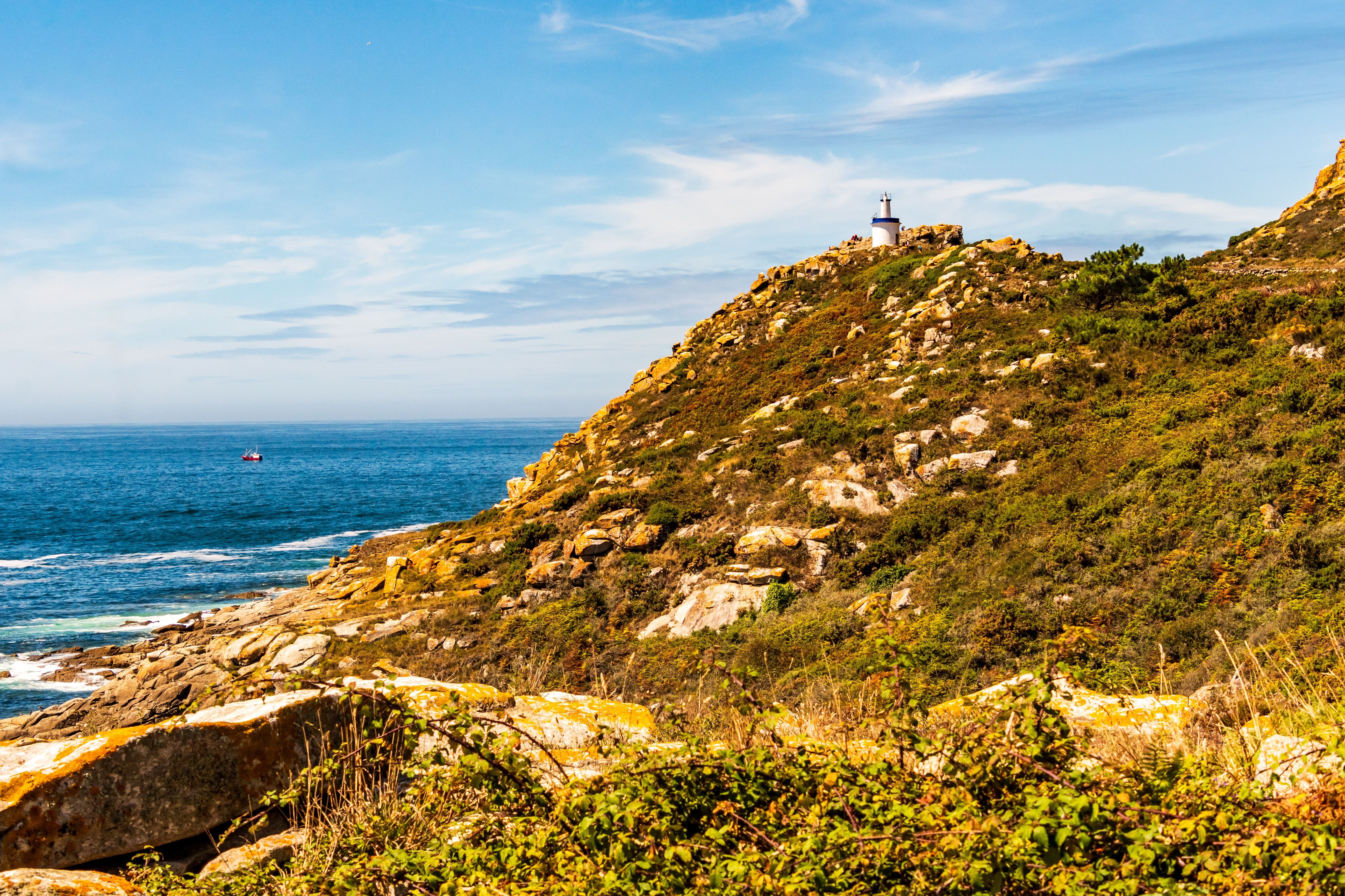 View of across rocky, scrub-covered cliffs towards a small lighthouse looking over the sea.